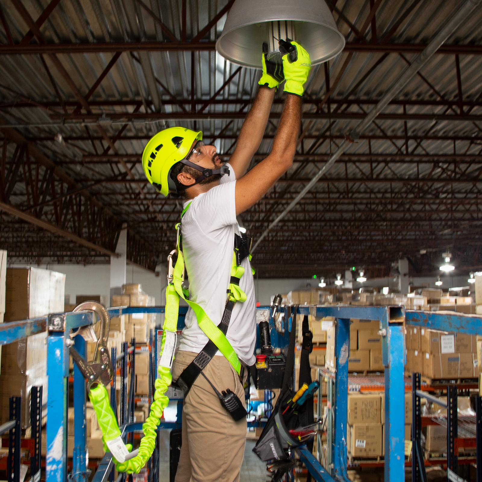 A man wearing the 1D ring JORESTECH fall protection safety body harness with grommets. He is standing on a lift while replacing light bulbs in a warehouse at 30 meters height. The man is attached to the structure of the lift by a lanyard hooked to the D ring of the JORESTECH harness and the metal structure of the lift.  There are rack, bins and brown card boxes bellow.