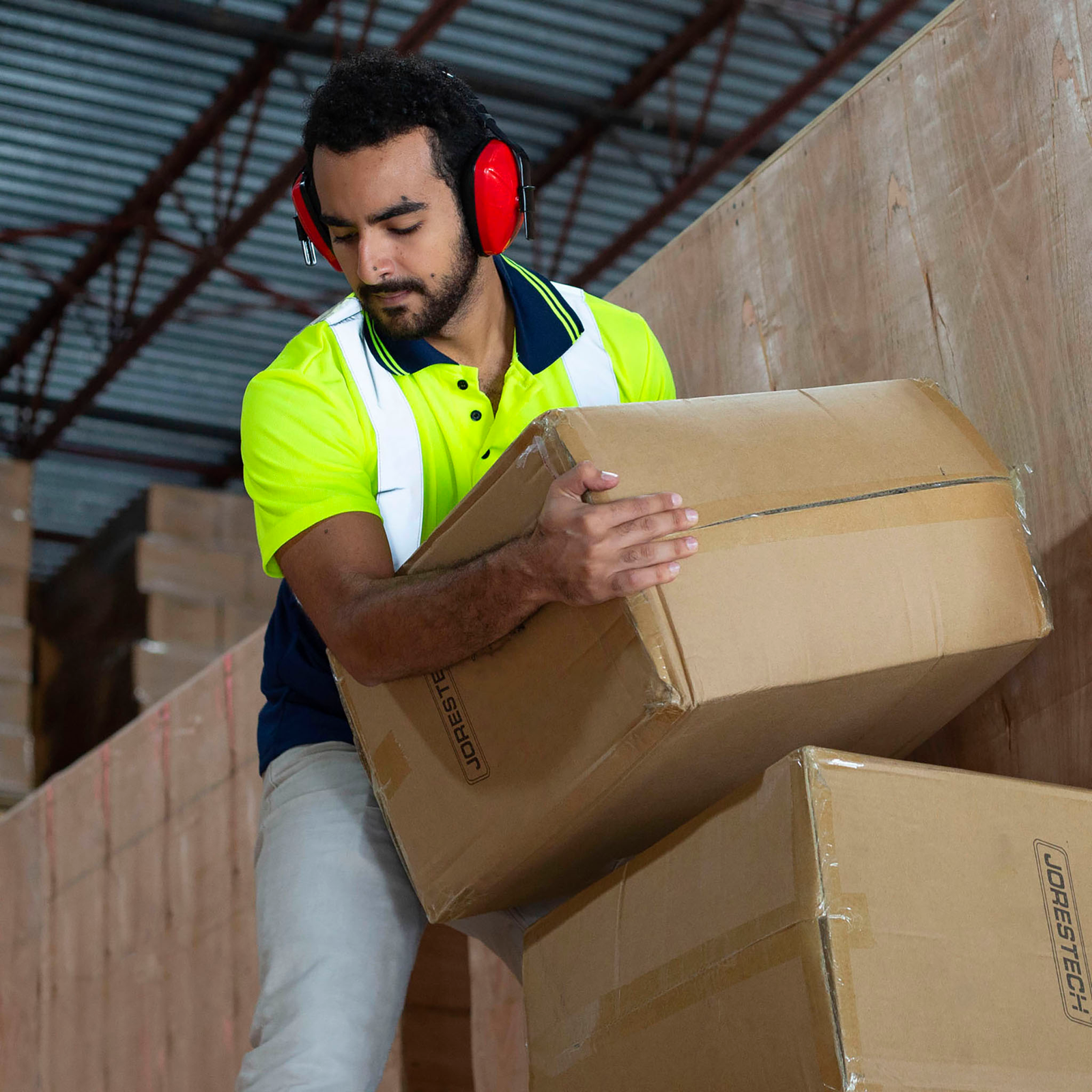 A man  is wearing red JORESTECH ear muffs for noise canceling while carrying boxes in a manufacturer