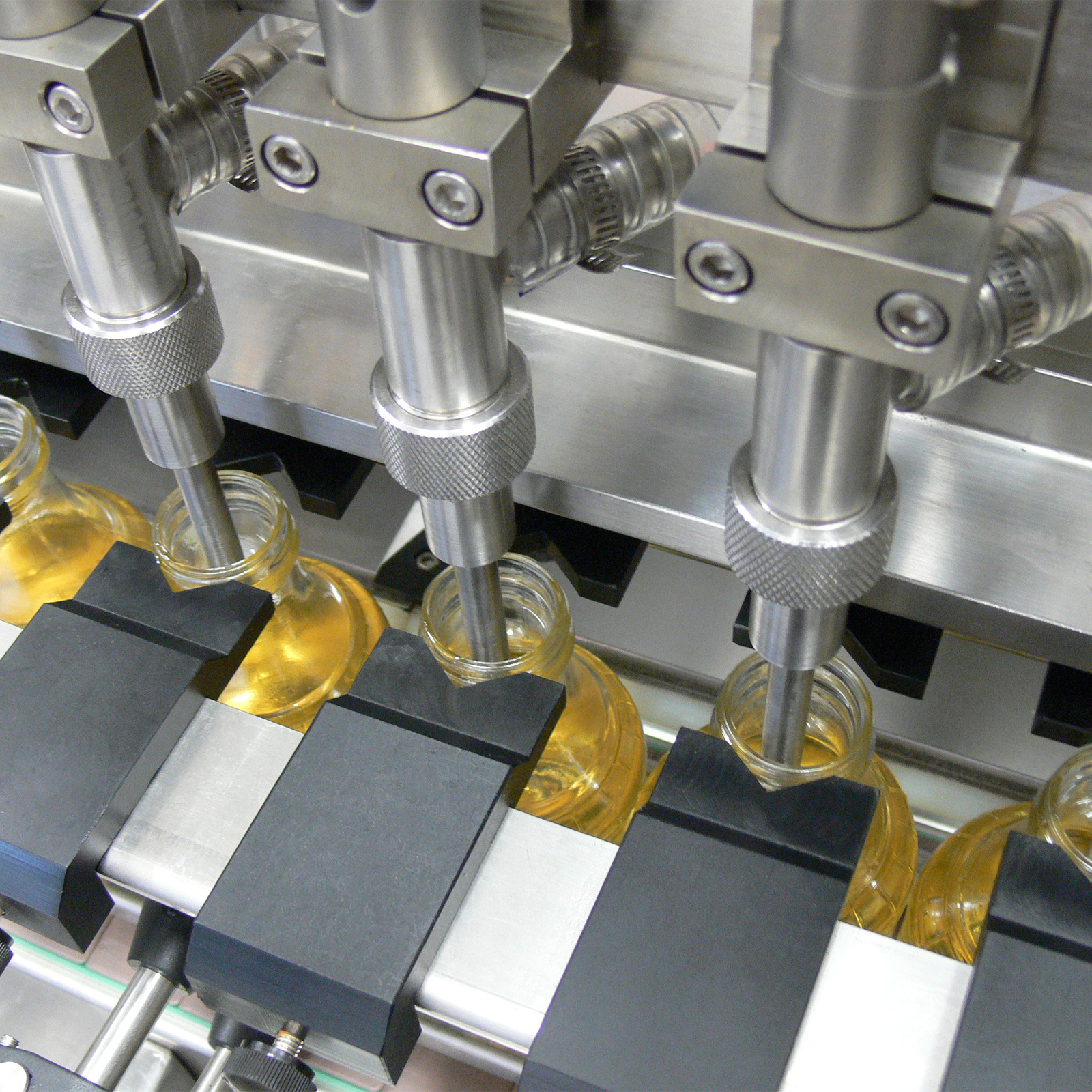closeup of the heads of the inline liquid filling system machine dispensing a yellow liquid into clear glass containers.