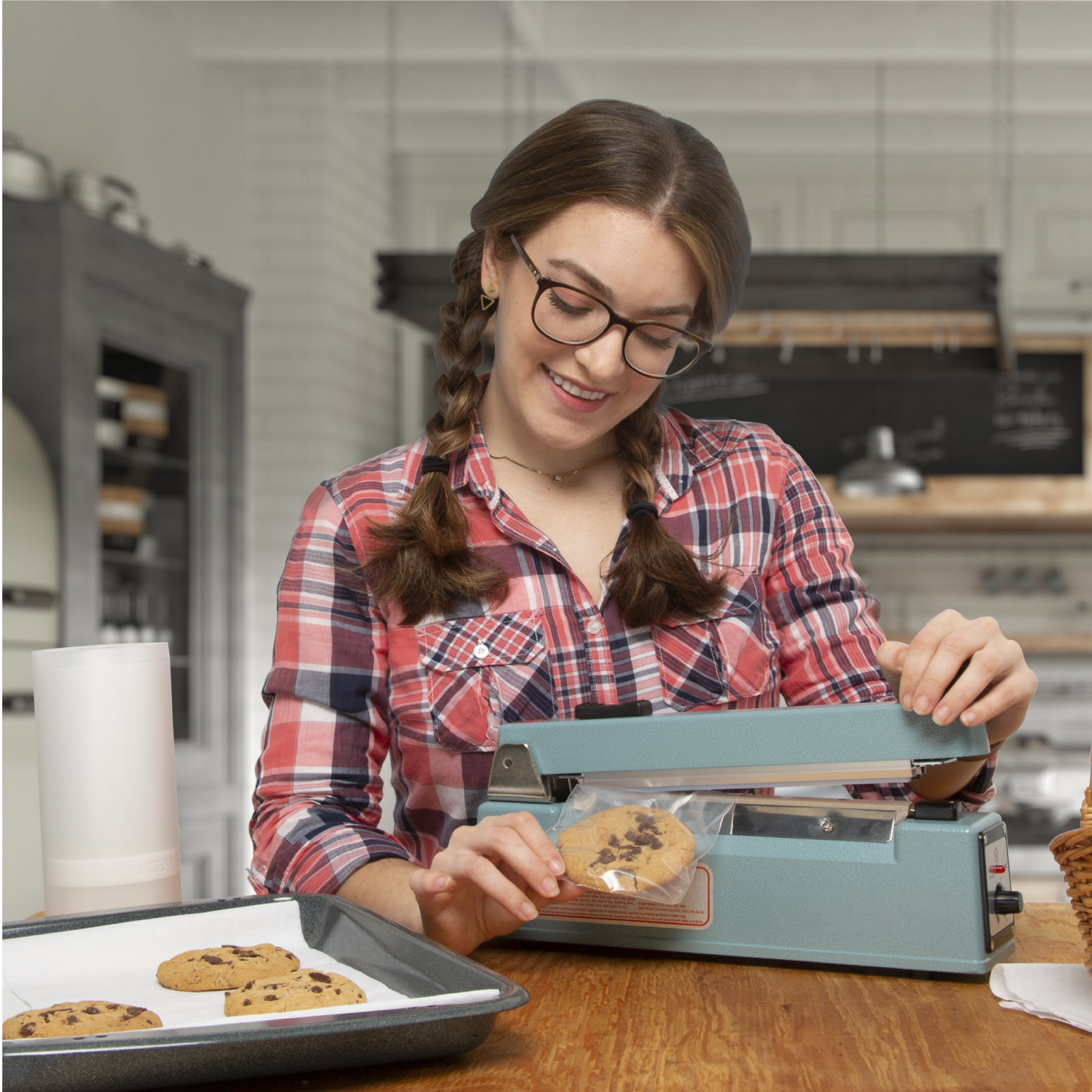 Young teen age girl in a kitchen. She is wearing a pink and blue plaid shirt and is sealing a batch of chocolate chip cookies in individual sealable plastic bags using the 8 inch manual impulse sealer