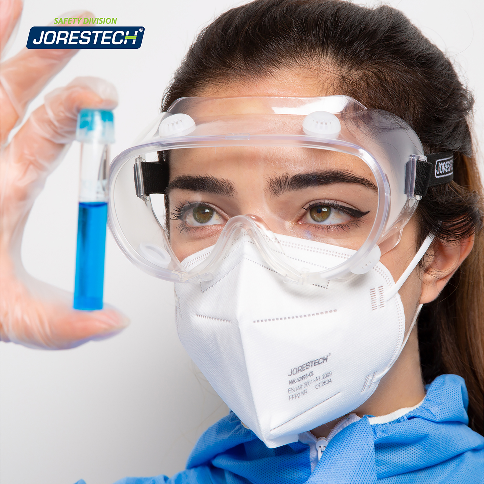 Woman wearing the Anti fog ventilated safety goggles and disposable clothing in a setting of a laboratory. She is inspecting a blue liquid inside a glass tube