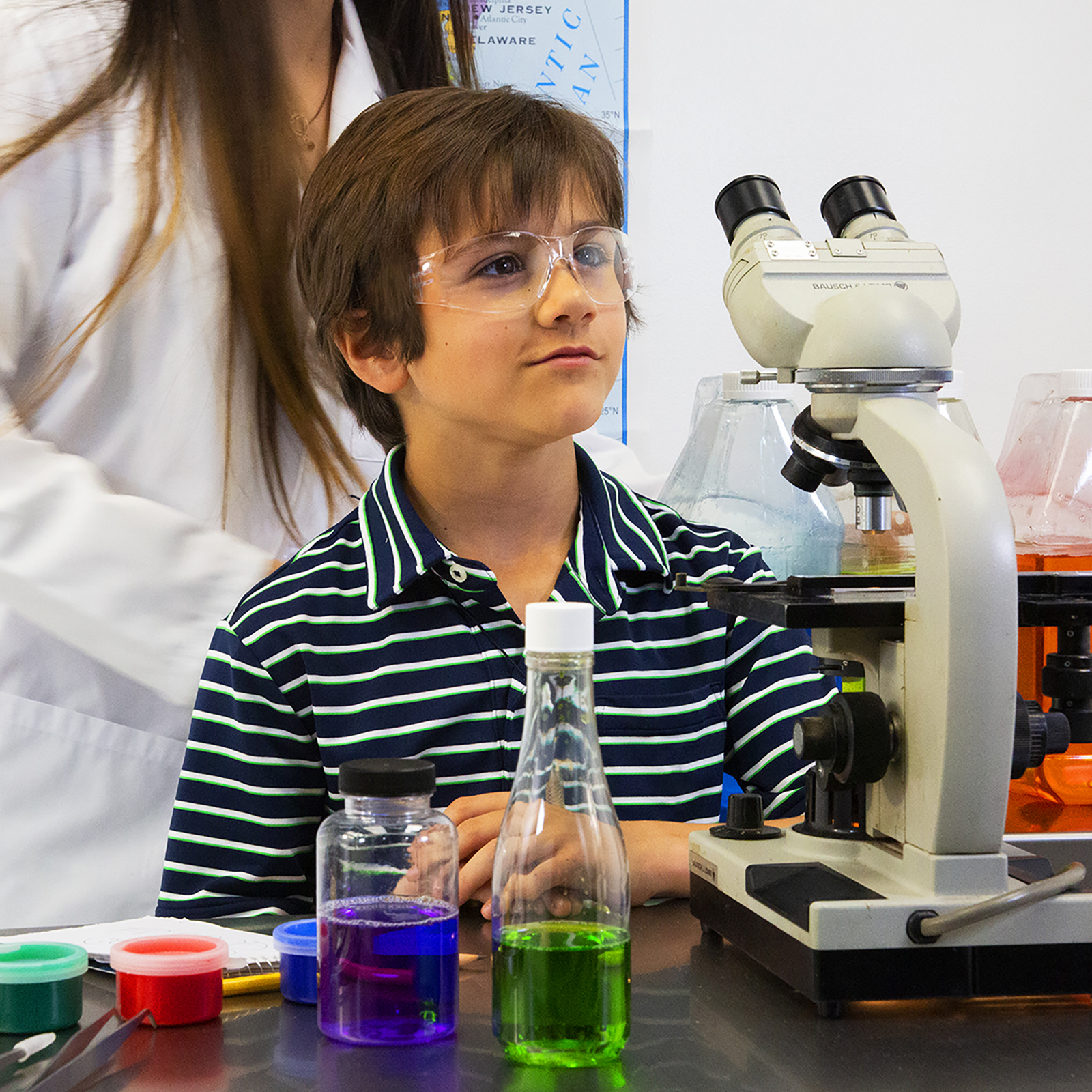 A boy wearing clear children's JORESTECH safety glasses for high impact in a lab setting. He is using a microscope and has a wide number of bottles with bright colored liquids to be examined. His teacher is in the background.