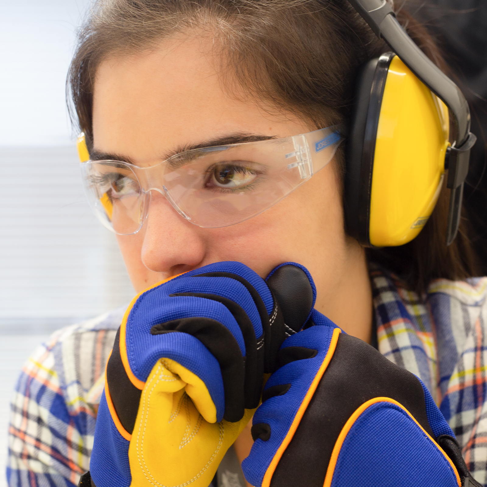 Young brown-haired woman wearing JORESTECH clear impact safety glasses, blue and yellow work gloves, and yellow earmuffs. 