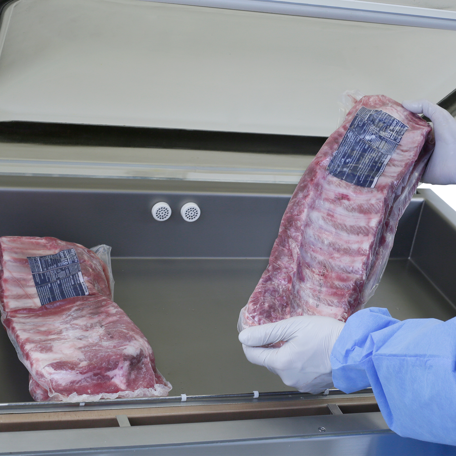 Person wearing protective disposable gloves sealing 2 large racks of ribs at the same time with a JORES TECHNOLOGIES® commercial single chamber vacuum sealer.   2 round vacuum vents are part of vacuum chamber