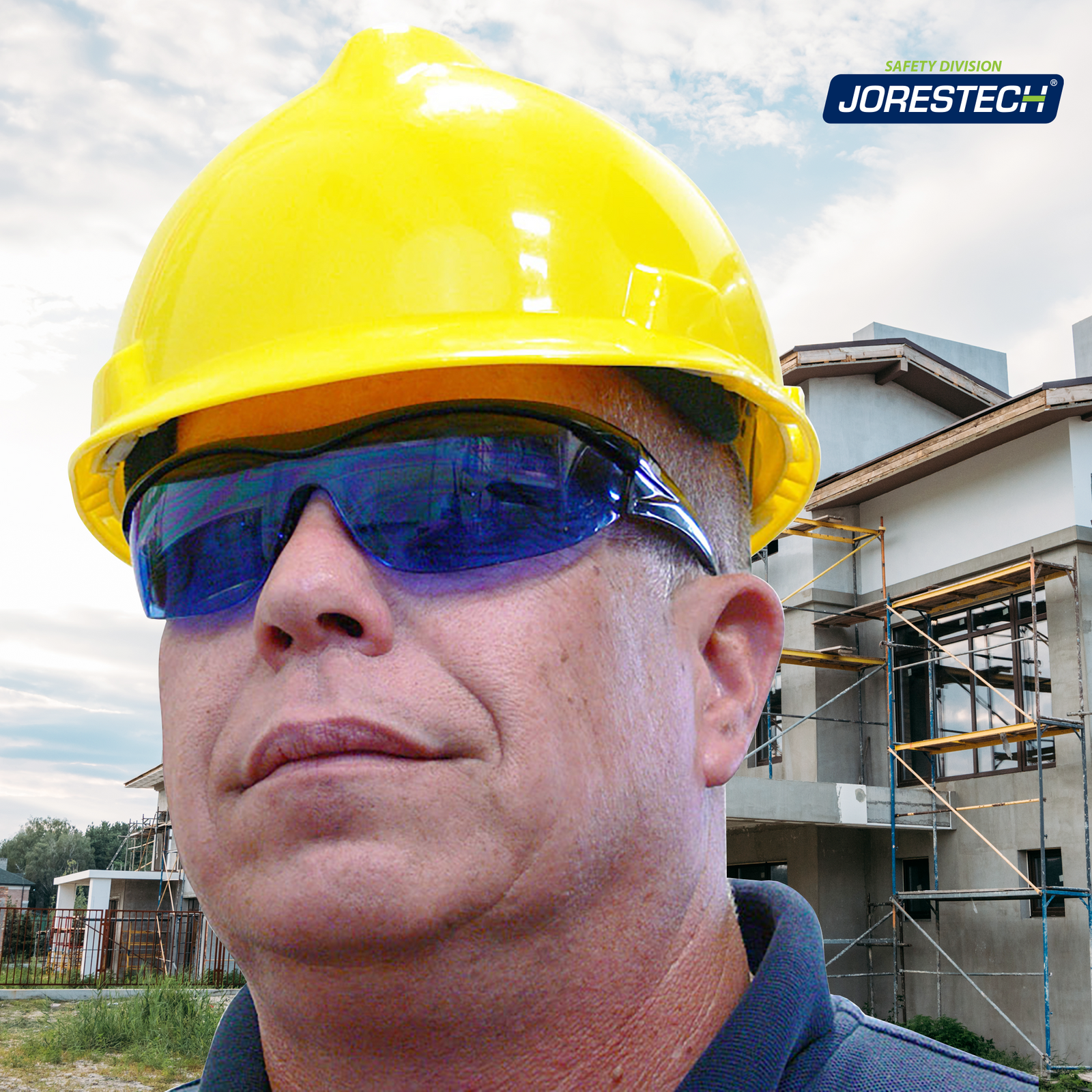 A man standing outside of a  construction of town houses. He is wearing the blue JORESTECH wraparound high impact safety glasses and a yellow cap style had hat. There are several scaffolds placed in front of the houses positioned to install windows and continue painting