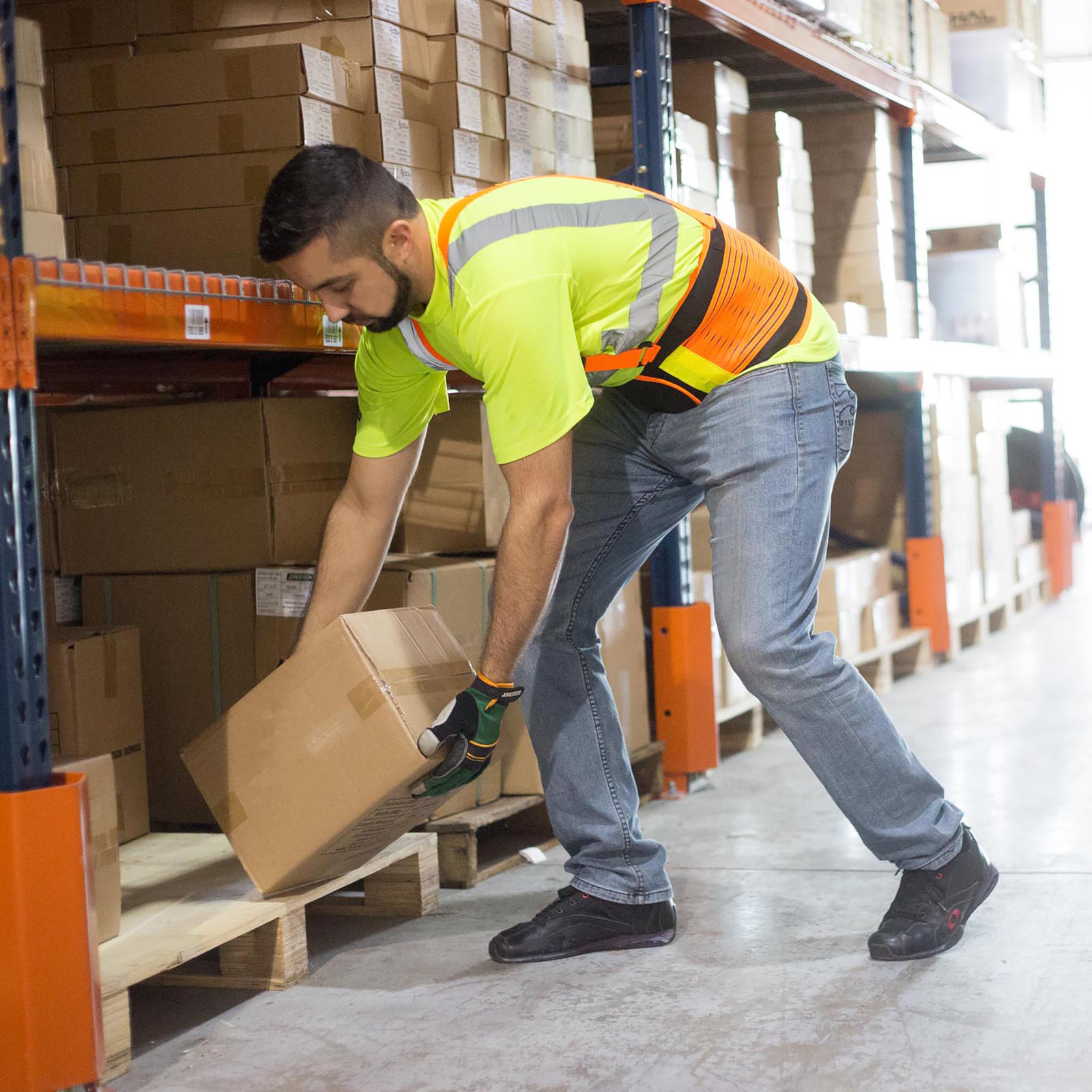 A worker wearing a high visibility adjustable heavy duty back support belt while organizing boxes inside storage racks in a warehouse
