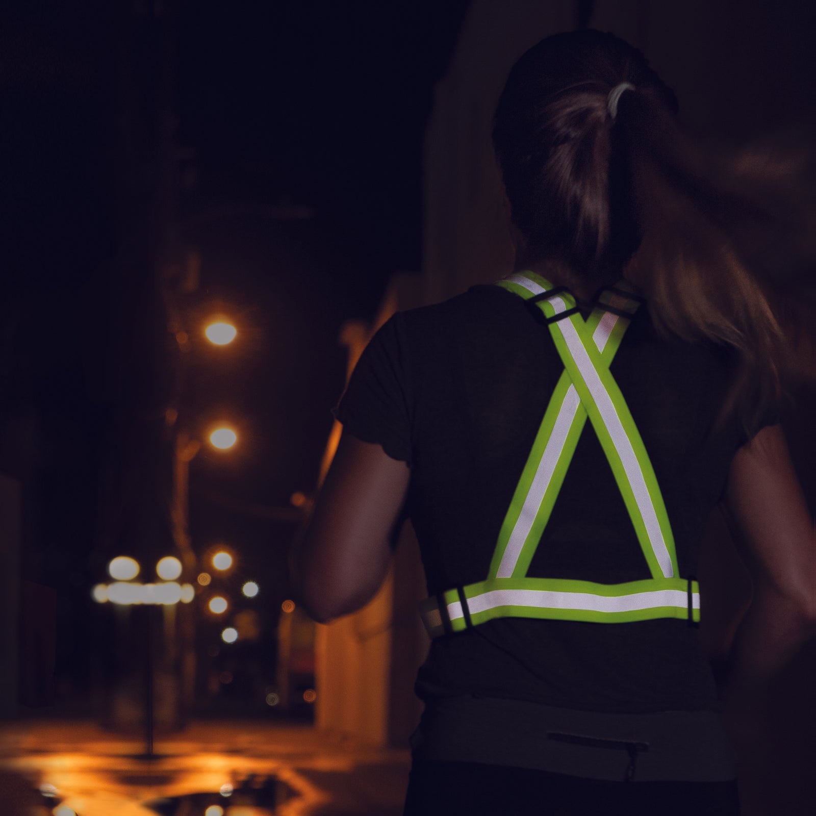 A woman wearing a lime hi vis JORESTECH adjustable safety suspenders while she is running or walking on the street at night time