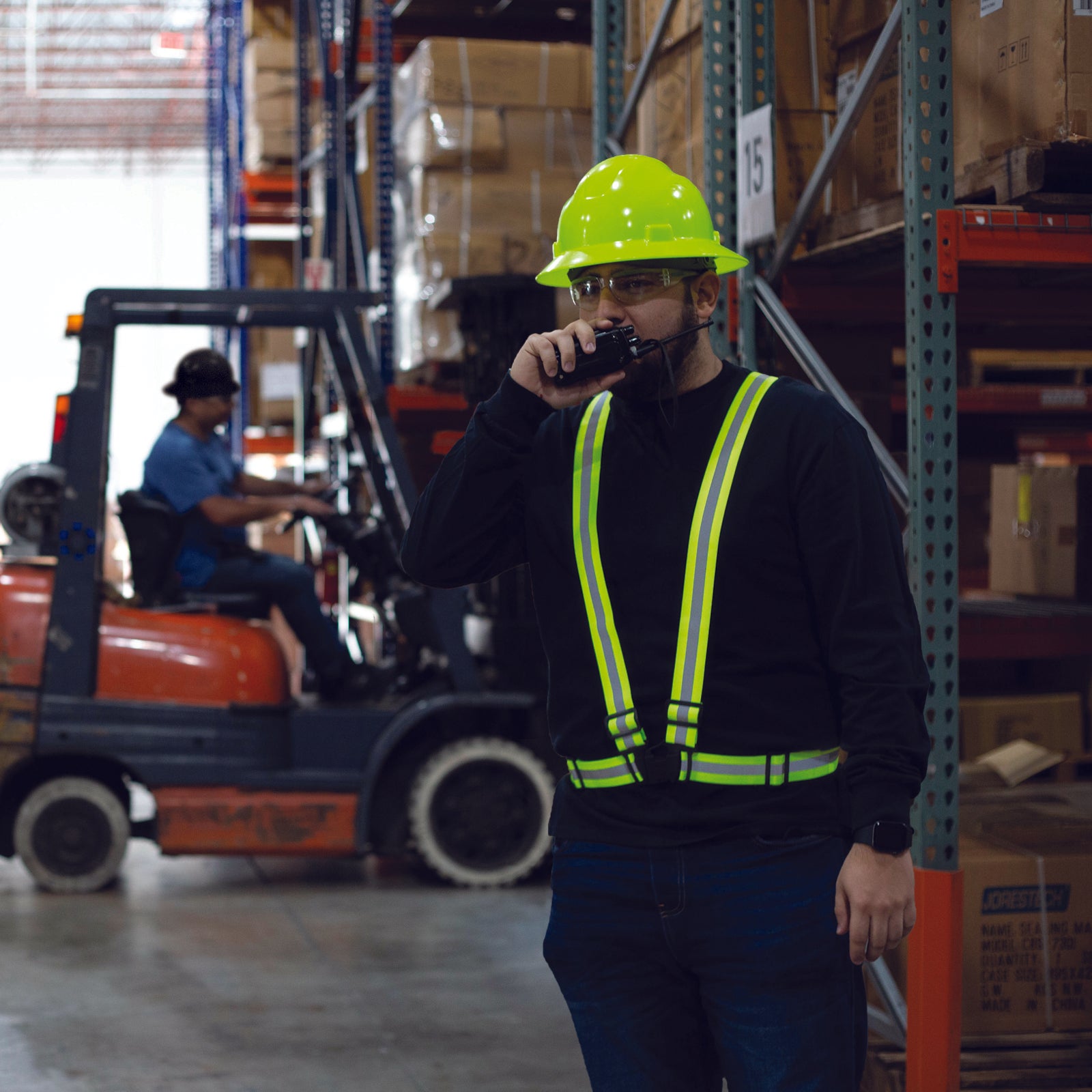 A worker wearing the lime JORESTECH safety suspenders inside a warehouse. There is a driver with a red forklift close by.