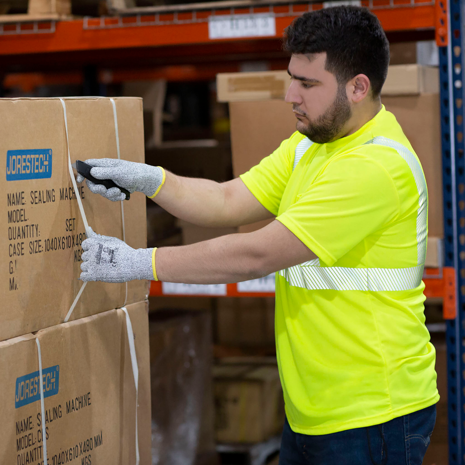 Image of a man wearing a JORESTECH yellow/lime safety shirt while removing strap form boxes while working in a warehouse