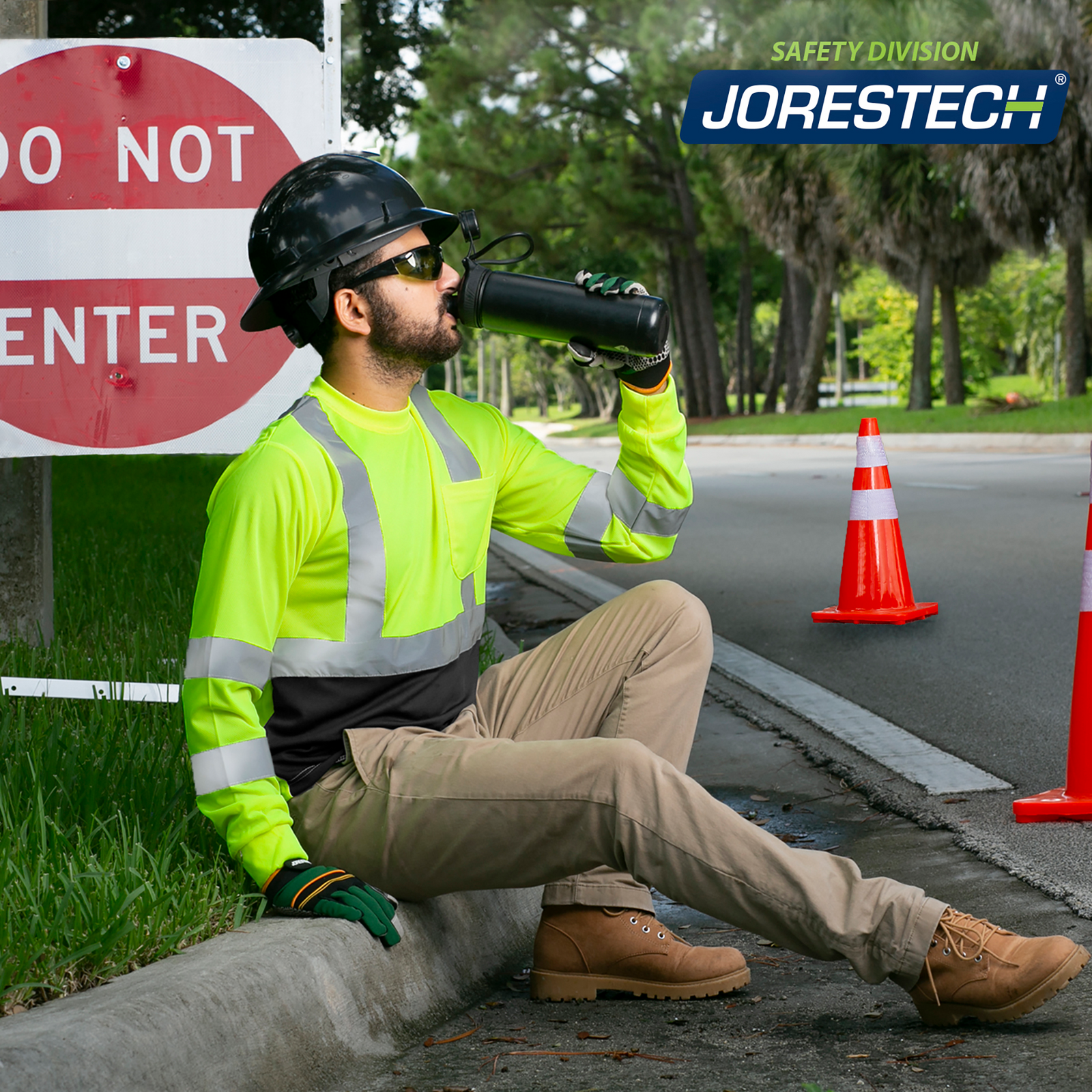 A worker wearing a JORESTECH hi vis safety shirt and a black hard hat while drinking water on the side of the road