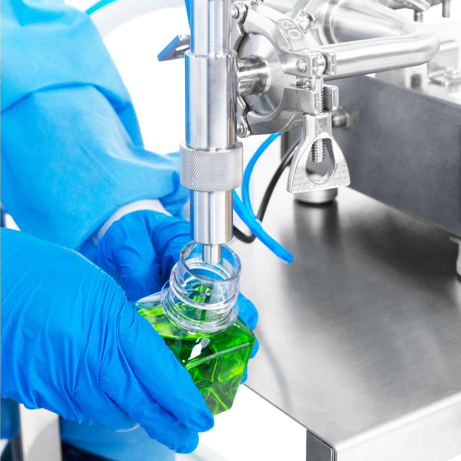 A person wearing nitrile safety gloves operating the Low Viscosity Piston Filler by JORES TECHNOLOGIES®. The worker is positioning a 100ml bottle below the tip of the non-drip nozzle to be filled with accuracy by the liquid filling machine.