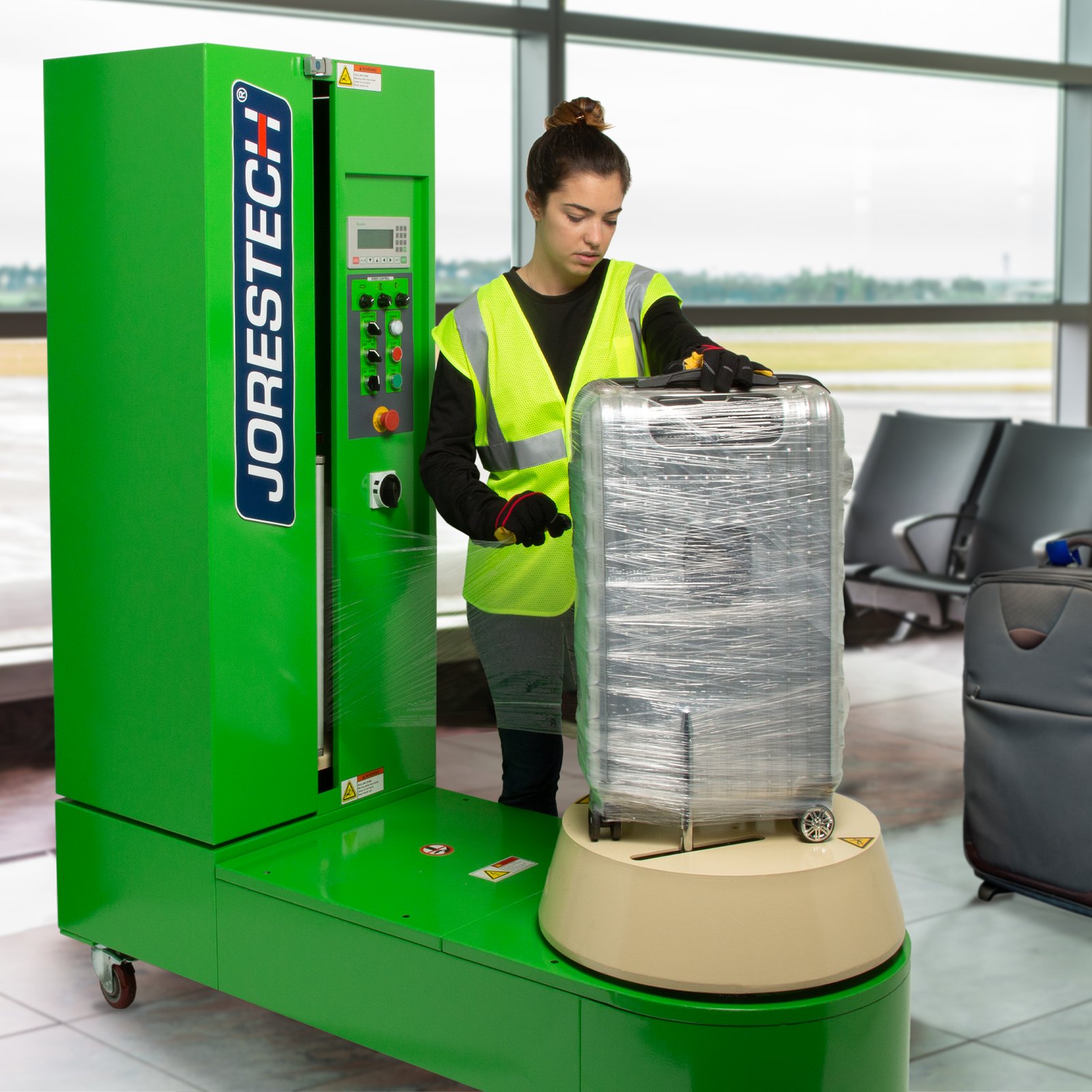 A woman wearing high visibility clothing in an airport using the JORES TECHNOLOGIES® Luggage stretch wrapping machine to wrap suitcases with stretch film.
