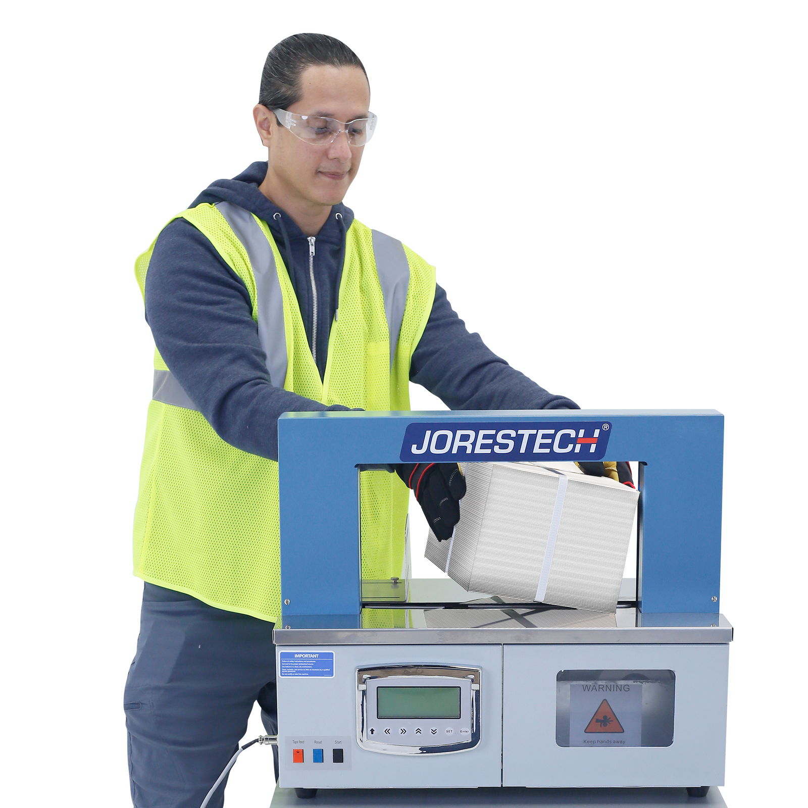 Man wearing a navy blue jacket and pants with a high-visibility yellow vest on top. He is using the JORES TECHNOLOGIES®  Paper banding system to apply and secure a band around a textured box.