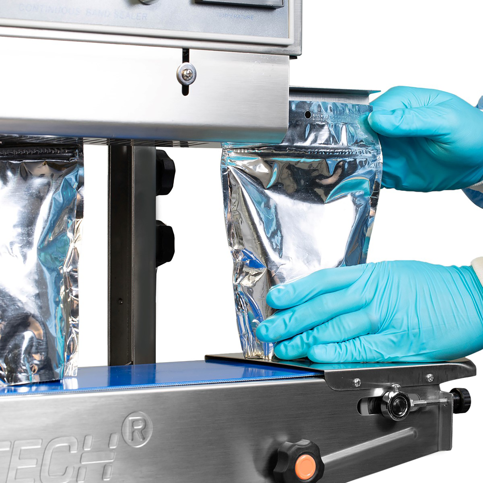 A person wearing protective clothing while inserting a silver plastic bag into the JORES TECHNOLOGIES® continuous band sealer. There are several bags on the blue revolving band being sealed.
