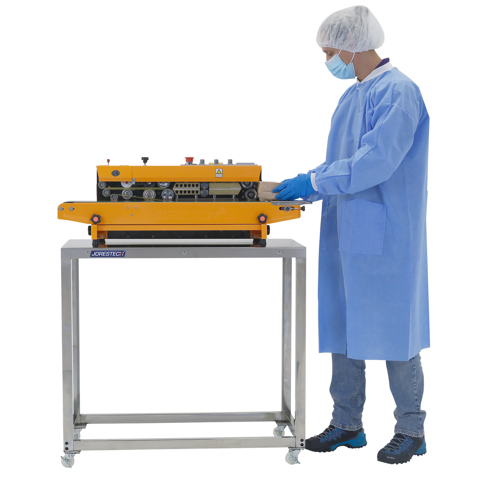 A person wearing disposable PPE and nitrile gloves using a continuos band sealer which is positioned on top of a JORES TECHNOLOGIES® Stainless steel prep work table