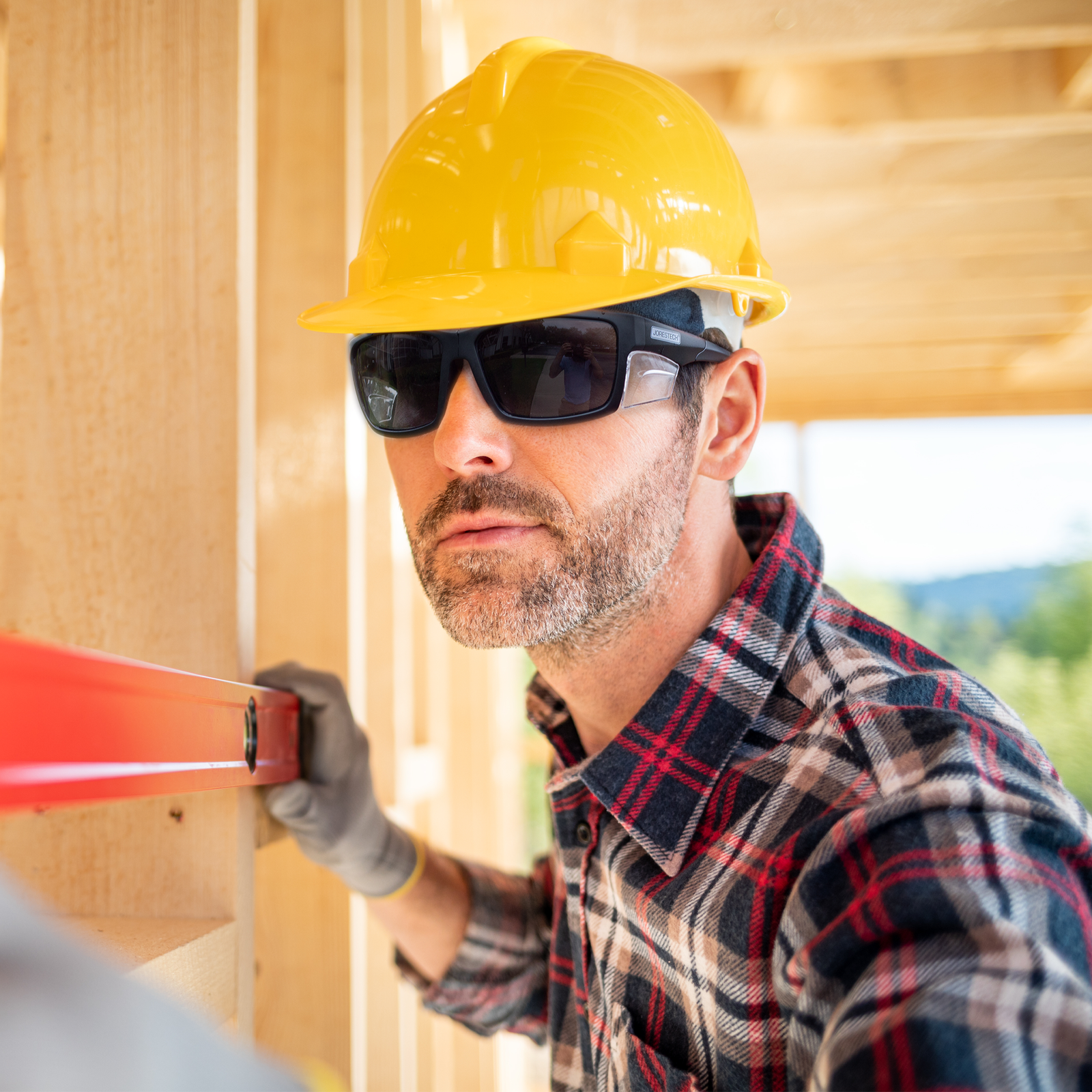 Image of a man wearing a yellow and the JORESTECH high impact sun safety glasses with side shields.. The man is checking the level of the wood structure of a house. The construction does not have walls or roof yet.