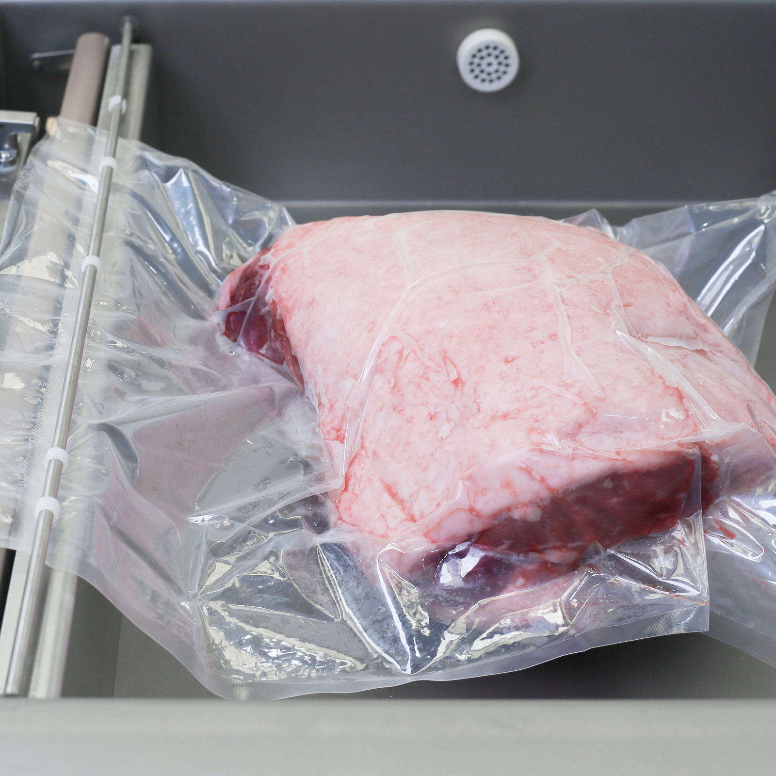 Vacuumed sealed bag with a large piece of meet inside. The bag is positioned inside chamber of the stainless steel JORES TECHNOLOGIES® tabletop commercial single chamber vacuum sealer with dual seal bar