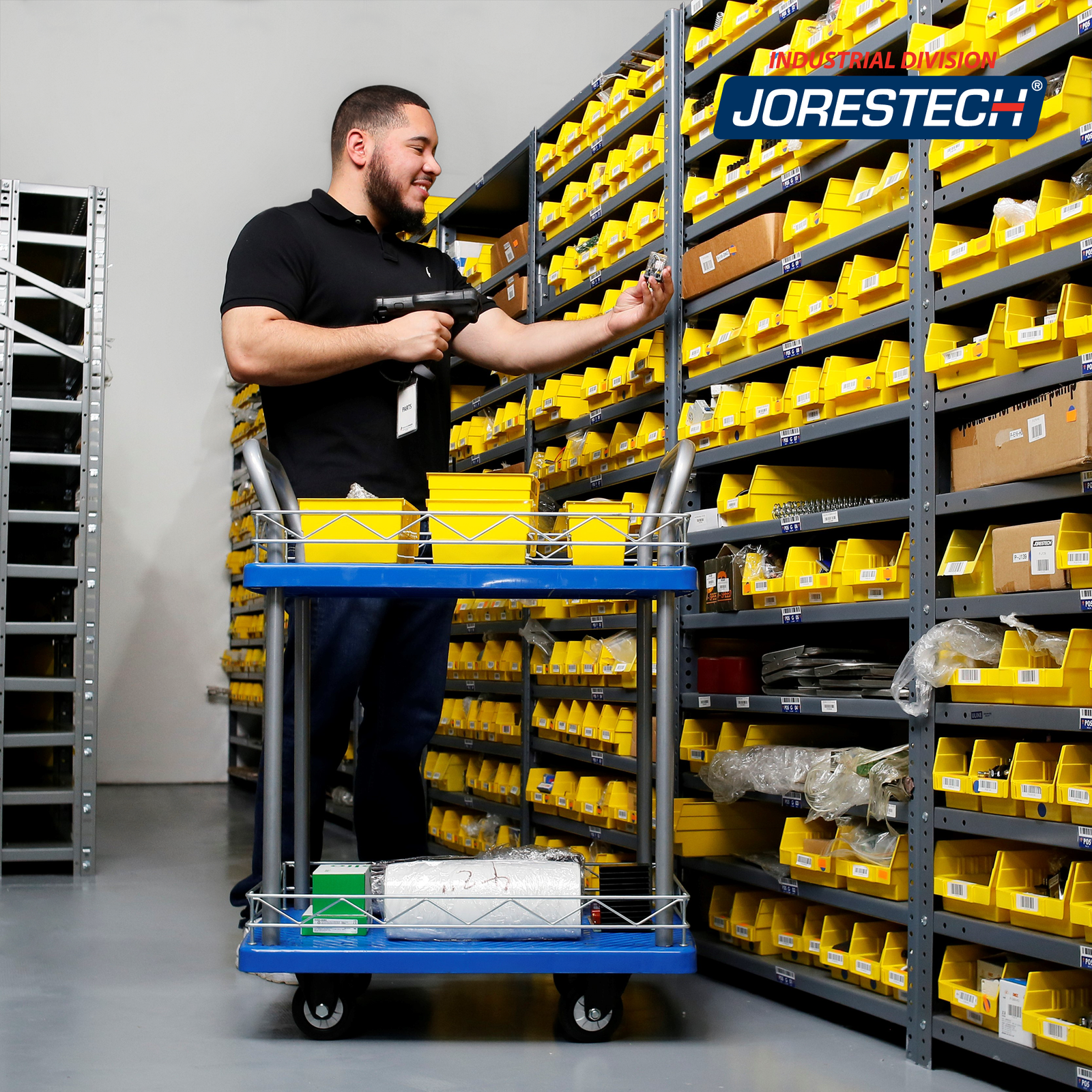 A man in a warehouse scanning some items and placing them on top of the dolly cart. He is using the JORES TECHNOLOGIES®  double platform utility push cart to roll the items around the workplace to reorganize