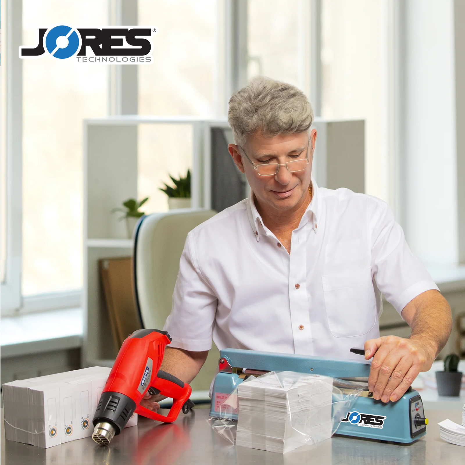 Man in an office setting sealing a pack of stationery paper with the manual impulse bag sealer with his left hand and he is holding a red heat gun with his right hand to shrink the film