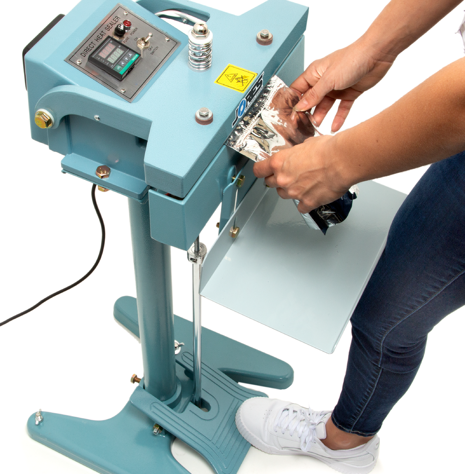Woman stepping on the foot pedal of the blue JORES TECHNOLOGIES® constant impulse bag sealer. She is sealing a silver plastic pouch bag.