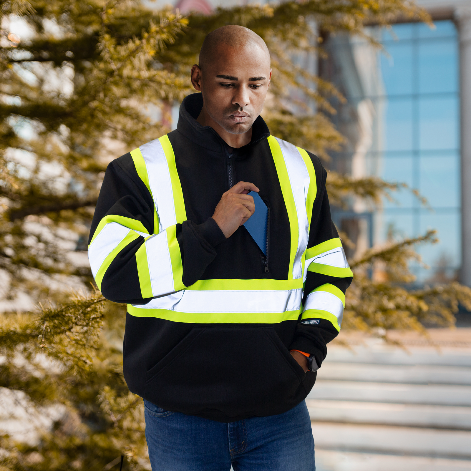 man storing his phone inside the chest pocket of the black safety sweater with reflective strips and contrasting yellow background