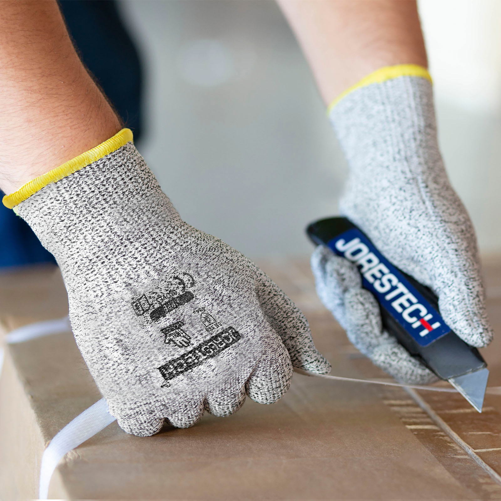 Hands of a person wearing the Jorestech cut resistant multi purpose safety work glove while cutting a strap with a box cutter