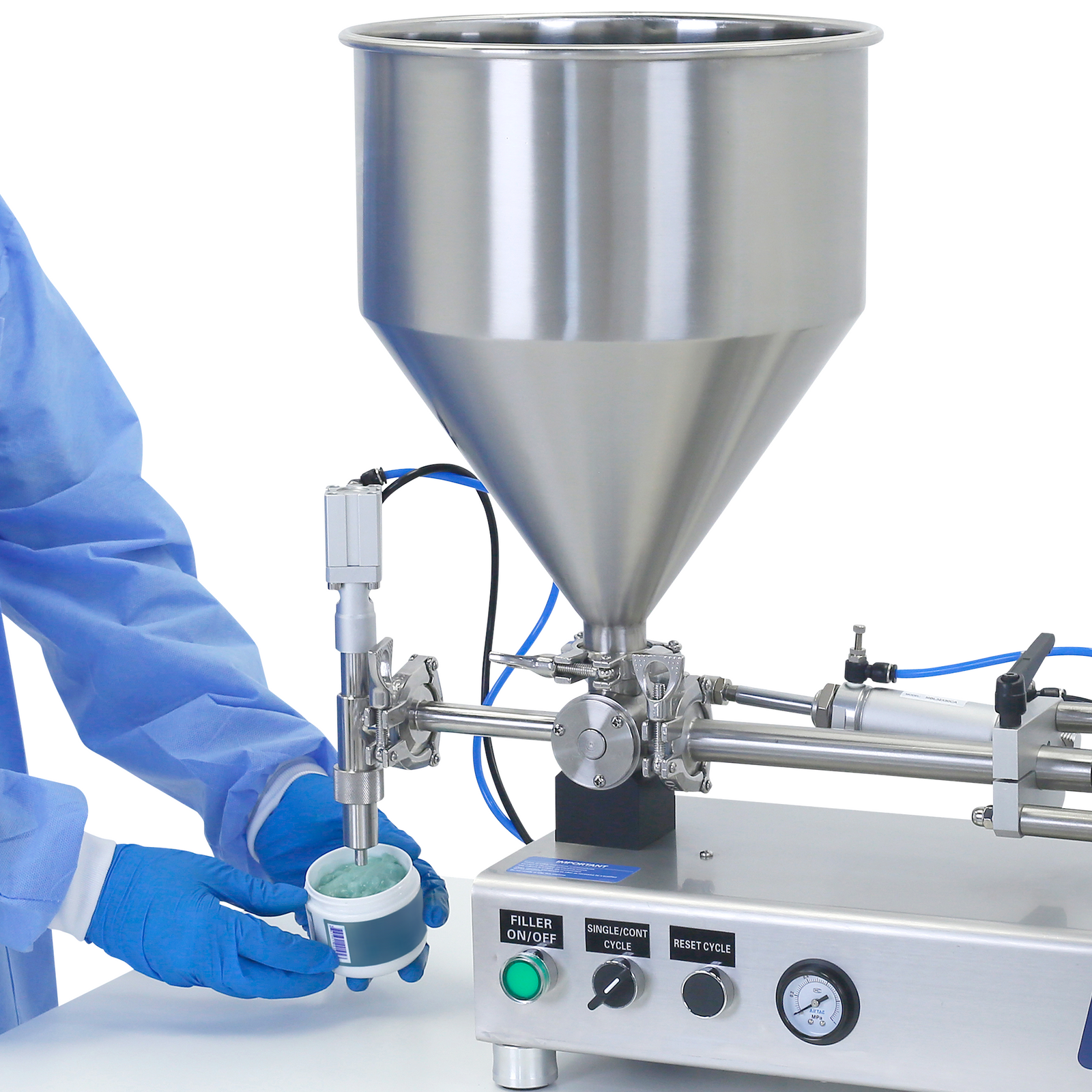 Close up of the hands of a person wearing PPE clothing while holding a container bellow the dispensing nozzle of the Piston filler for high viscosities. The container in been filled with a blue gel and the stainless steel piston filler in places on top of a working table