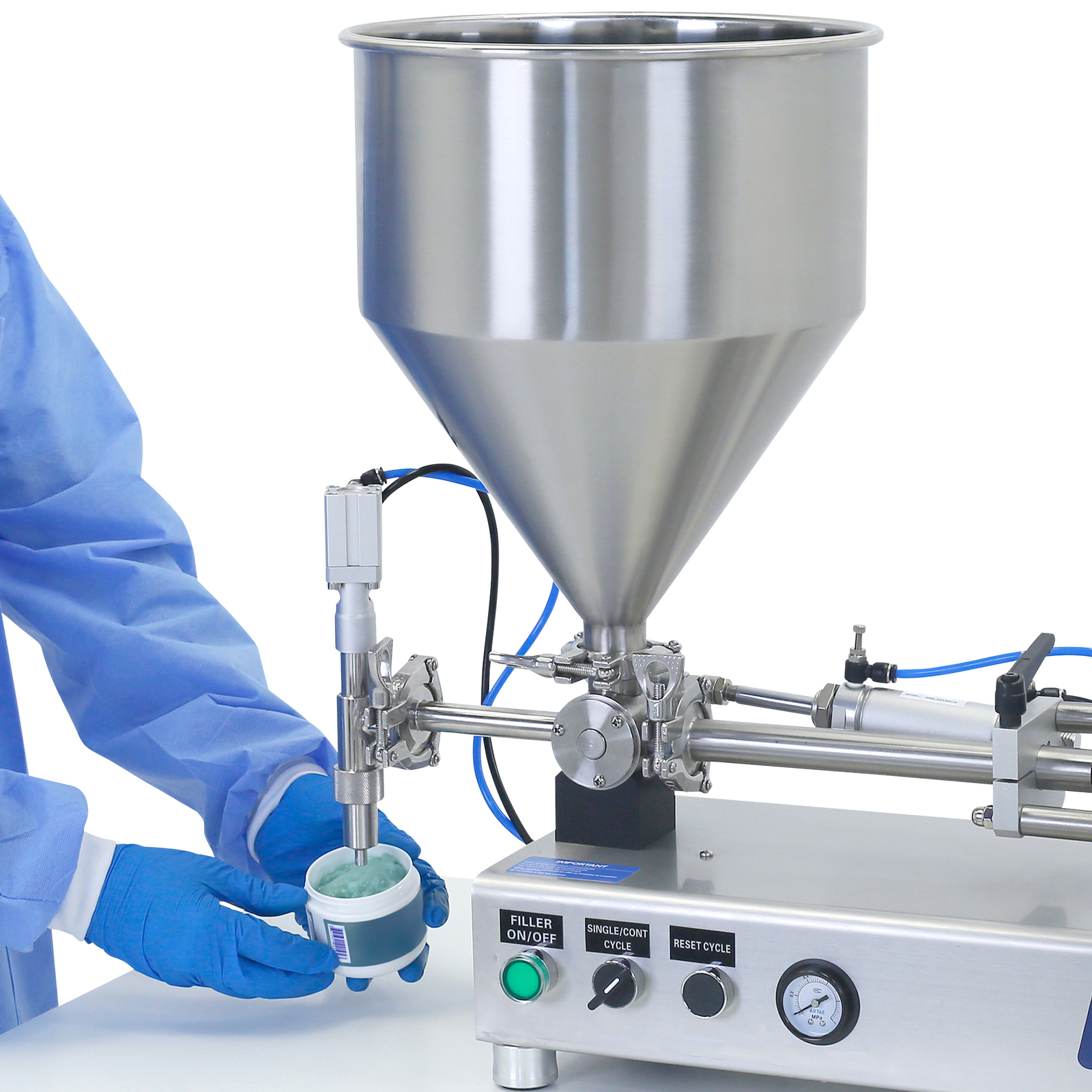 the hands of a person wearing PPE clothing while holding a container bellow the dispensing nozzle of the Piston filler for high viscosities. The small container in been filled with a blue gel and the stainless steel piston filler in places on top of a working table
