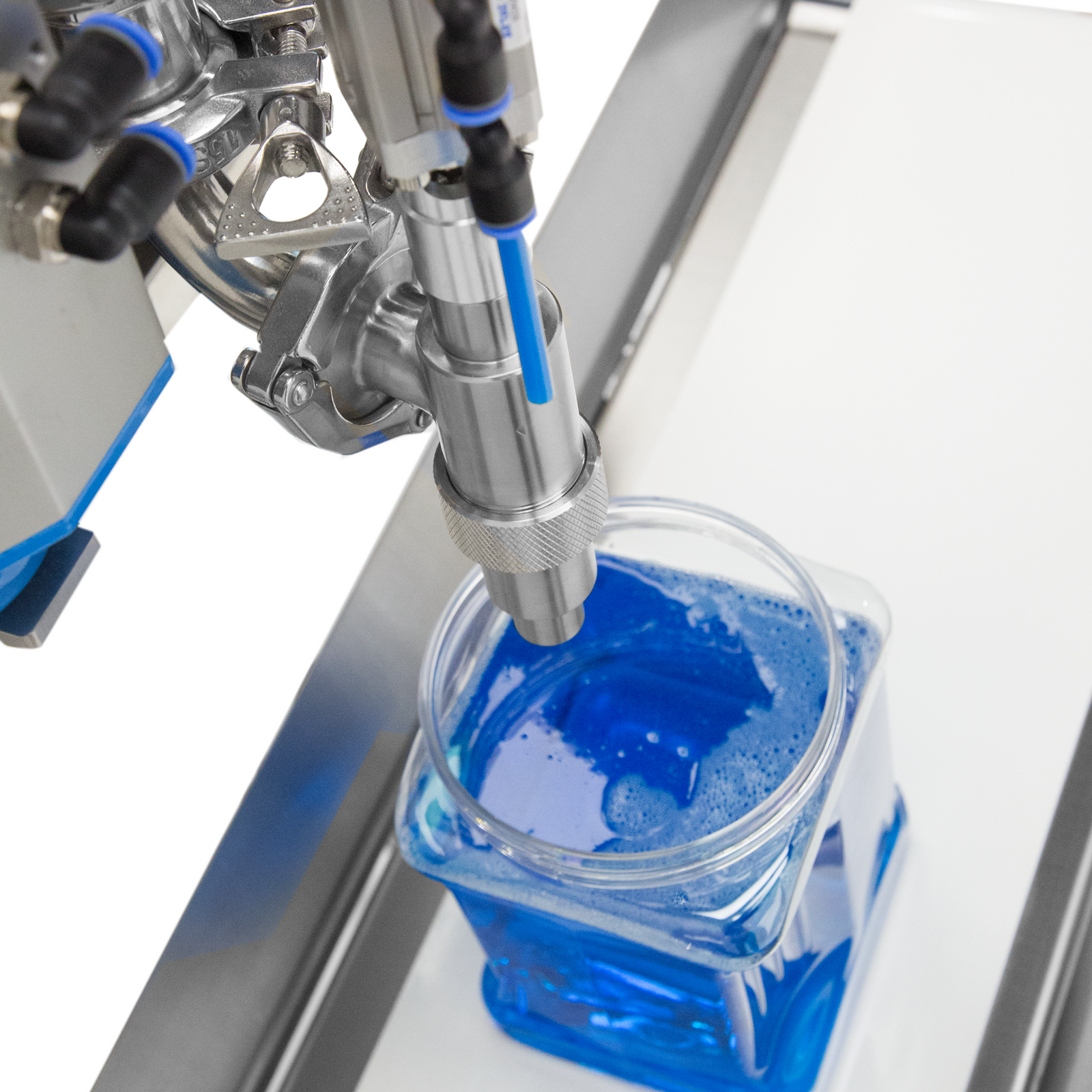 Closeup of the nozzle of the paste piston filler. There is a clear container that has been filled with blue liquid with the machine