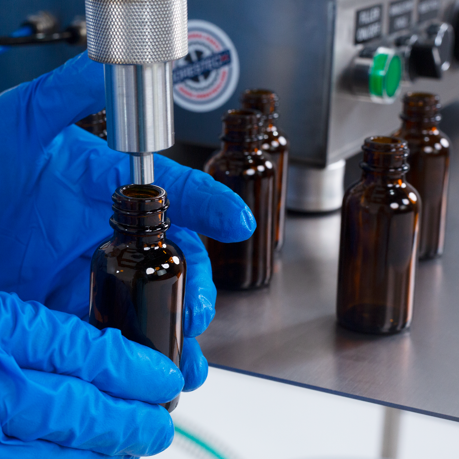 An worker wearing nitrile safety gloves operating the low viscosity table top Piston Filler for 50 ml. Is positioning small bottles below the  tip of the non-drip nozzle to be filled with accuracy by the liquid filling machine by JORES TECHNOLOGIES®