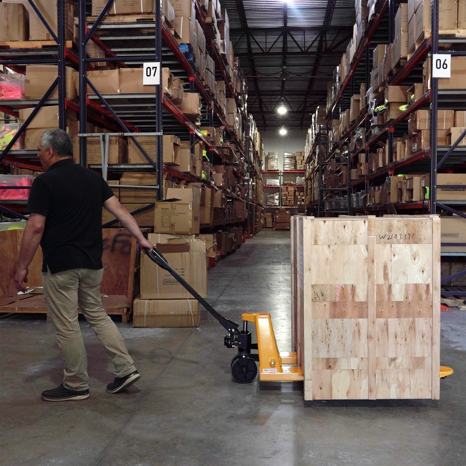 A worker pulling a yellow and black JORES TECHNOLOGIES® pallet jack truck in a warehouse filled wit boxes.. The pallet jack has several large heavy wood box on top