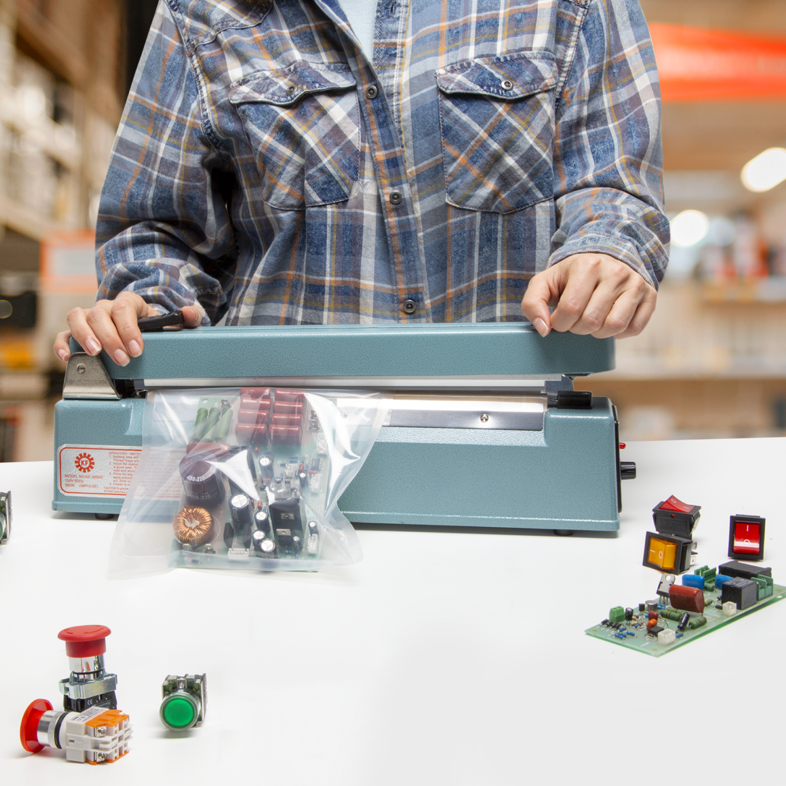 Person wearing a blue plaid shirt, she is sealing a bag with electronics inside using the 12 inch manual impulse sealer