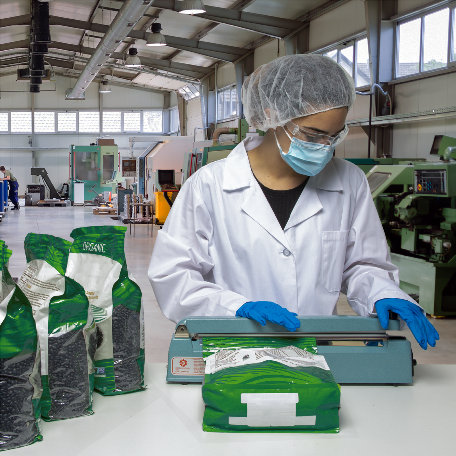 Woman in a factory setting wearing personal protection equipment and sealing several large sealable bags of organic black beans with the manual impulse sealer. 