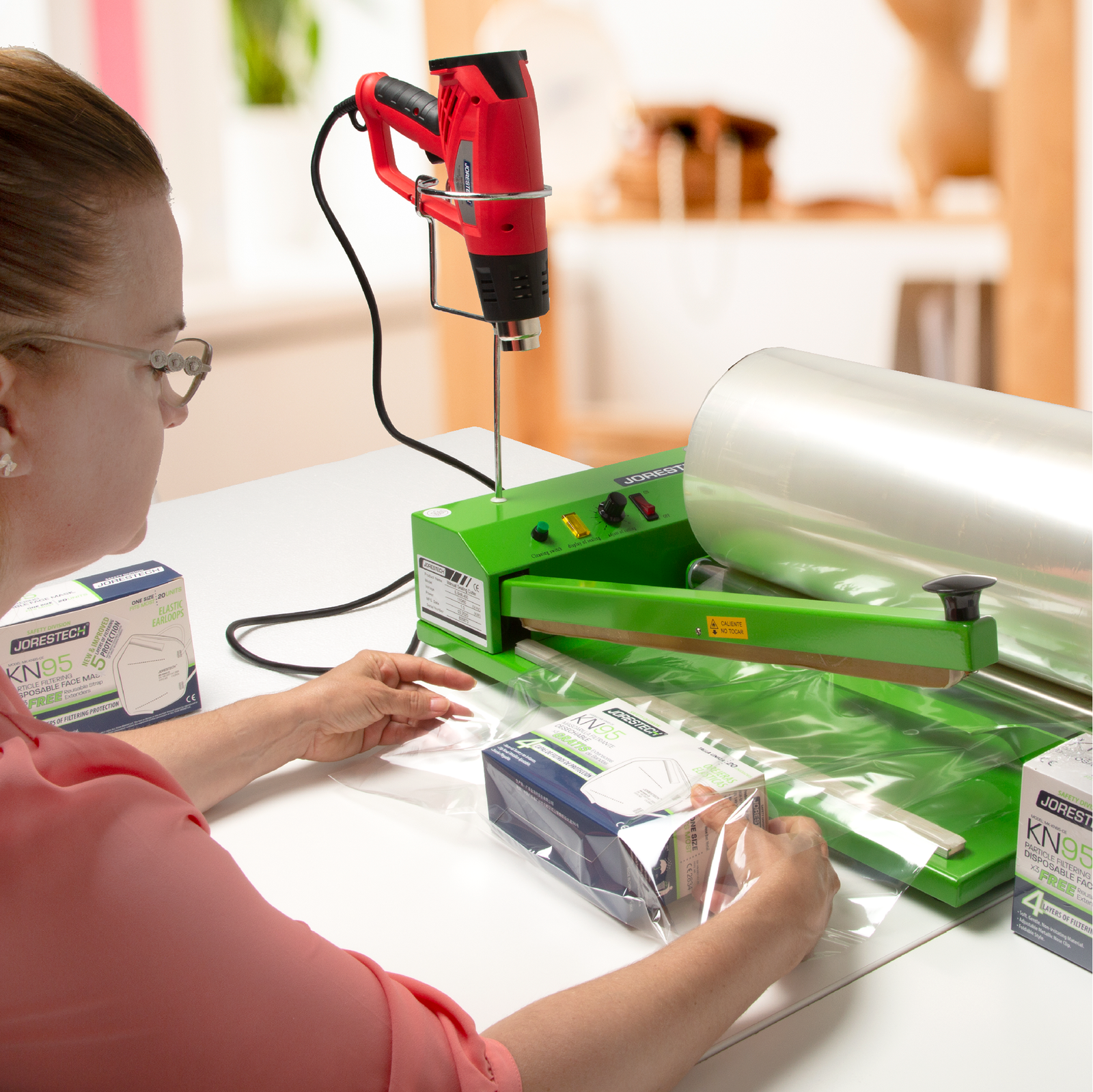 A woman in a pink shirt is using her shrink packaging unit to seal boxes of face masks. A heat shrink gun is mounted on the machine’s shrink gun holder and a roll of film rests on the roll holder bars. 