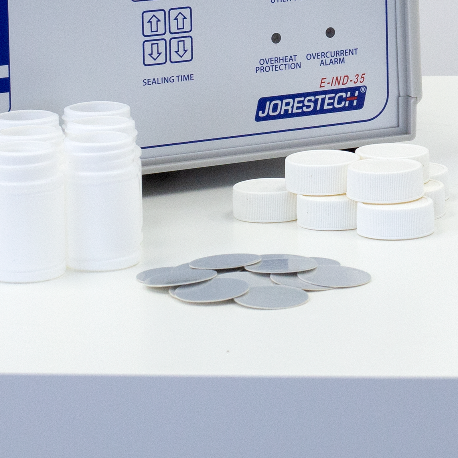 Image shows several container, several caps and several 2 piece induction liners on top of a table and a manual induction sealer in the background.