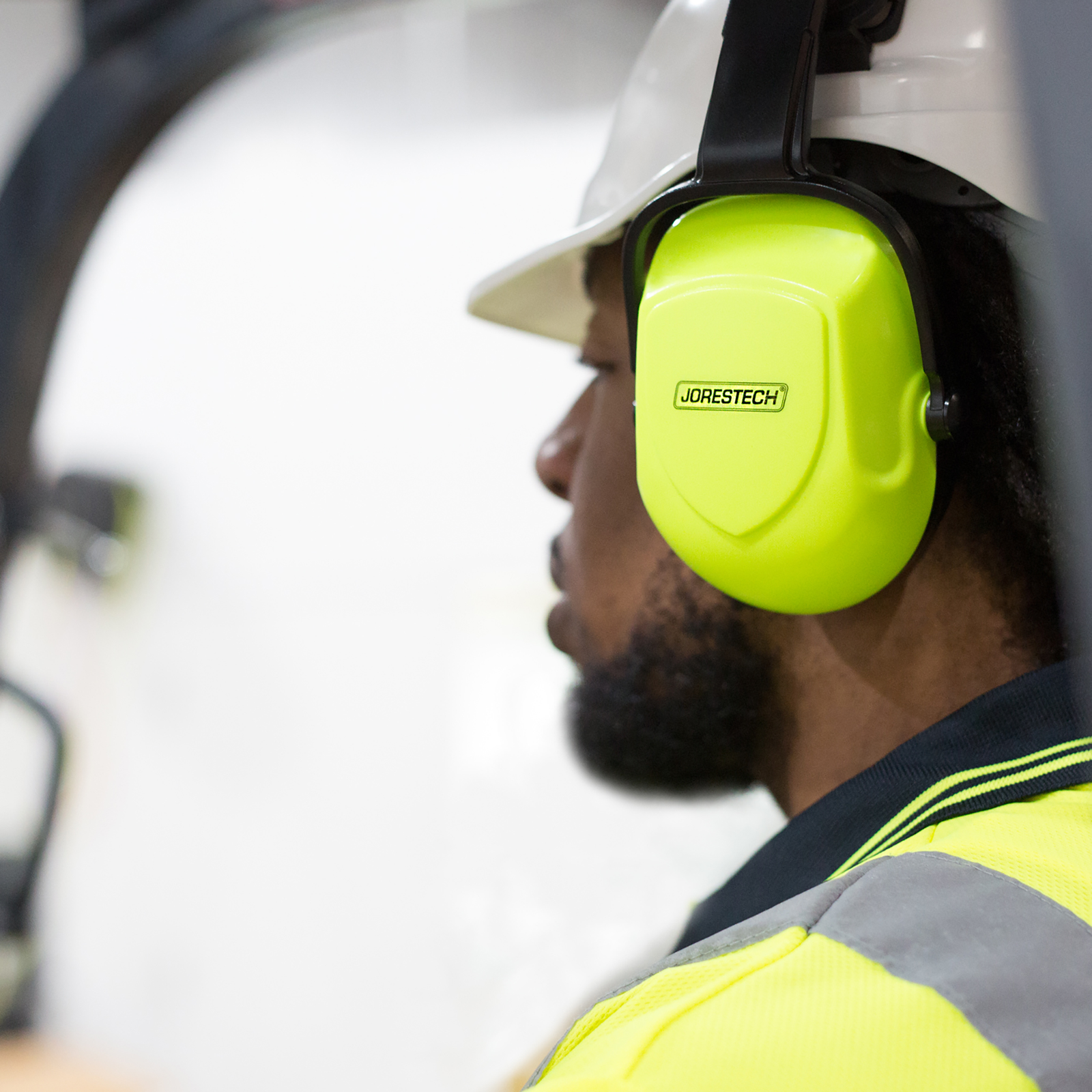 A worker wearing a white hard hat with the bright lime JORESTECH noise cancelling ear muffs installed