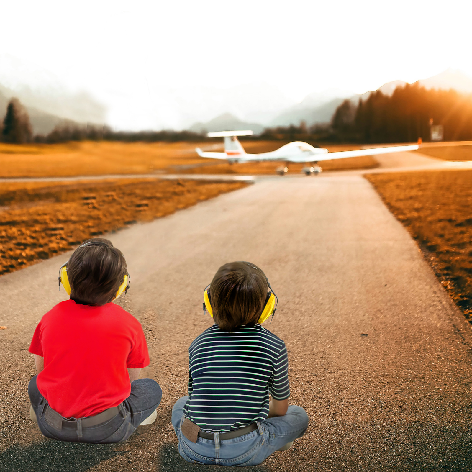 2 kids are sitting on the floor wearing yellow ear muffs while there is a airplane getting ready to depart