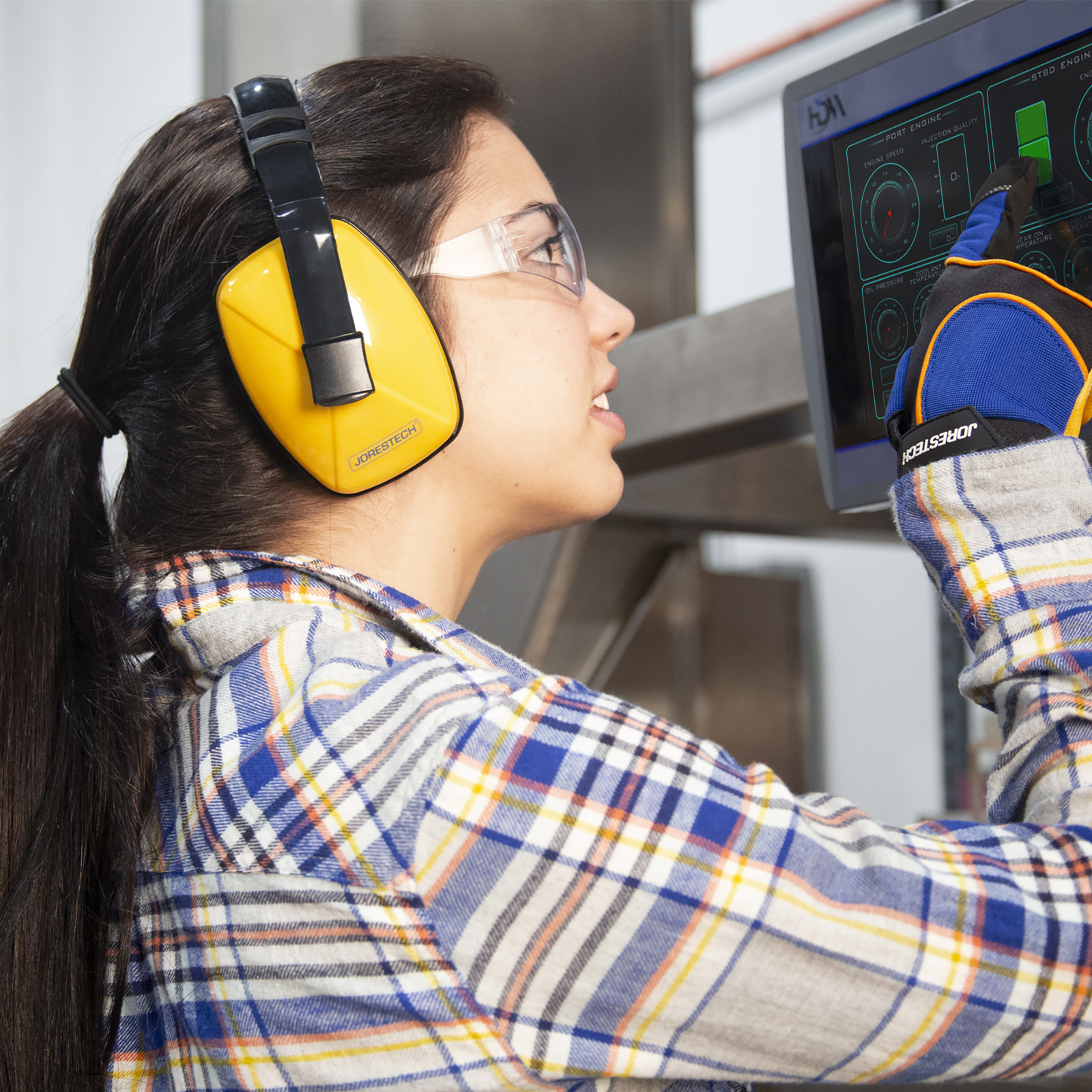 A woman wearing safety eye glasses, gloves and JORESTECH hearing protection ear muffs while operating a large loud machine