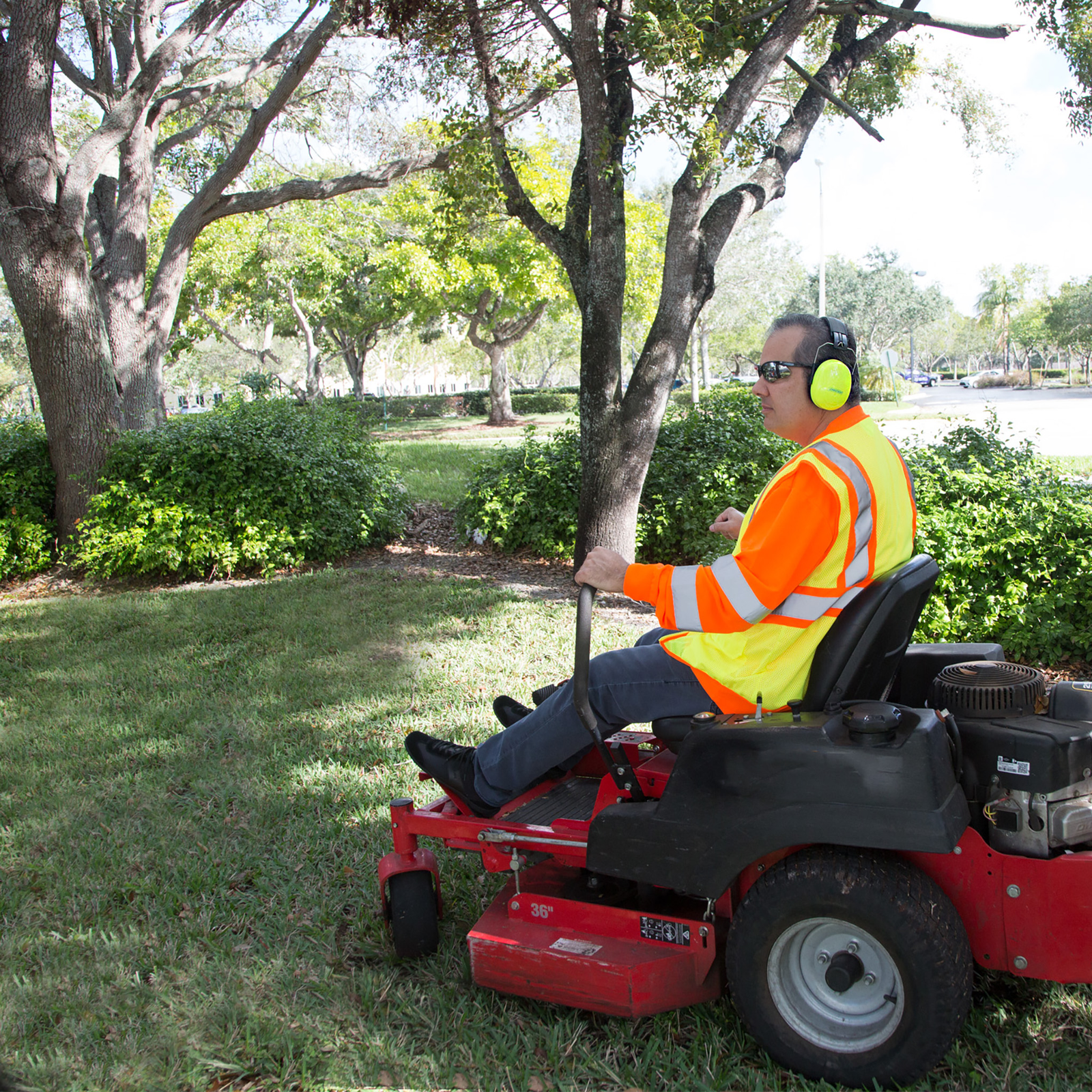 A garthener wearing a high visibility reflective vest and protective ear muffs while he is mowing the lawn with a red truck