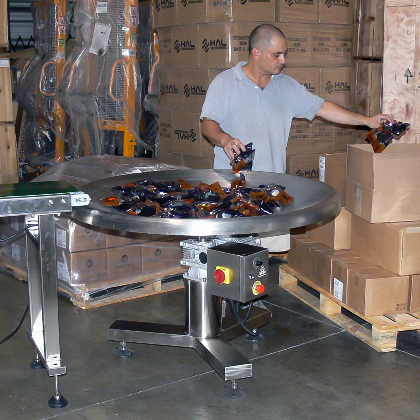 man wearing grey shirt moving packages from accumulating rotary table into a box with pallets of boxes in the background 