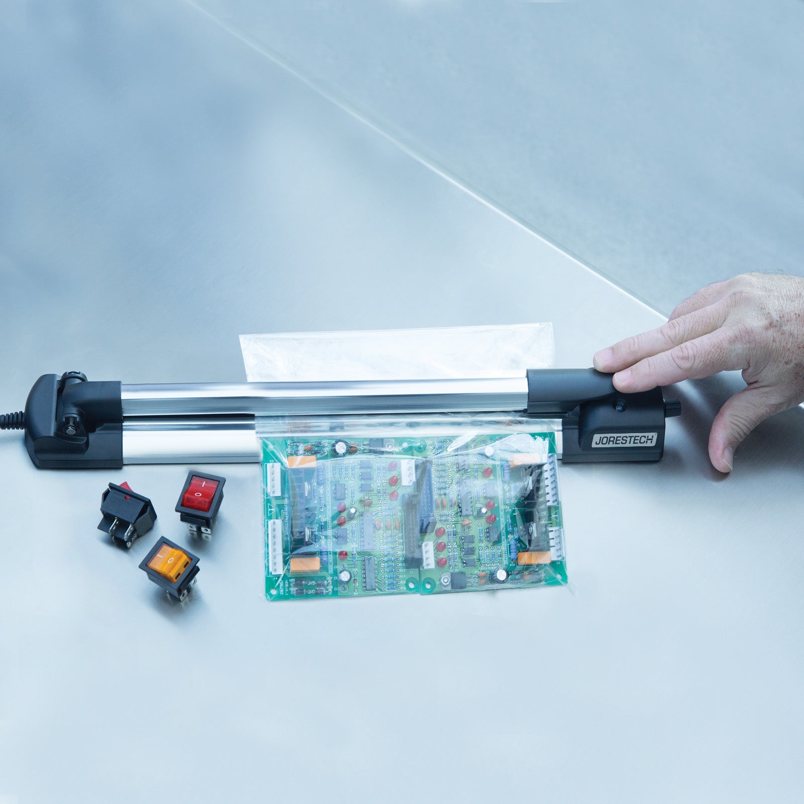 Hand of a man pressing the jaw of a Slim JORESTECH Manual Bag Sealer down to activate machine to seal a bag with a mother board and other parts
