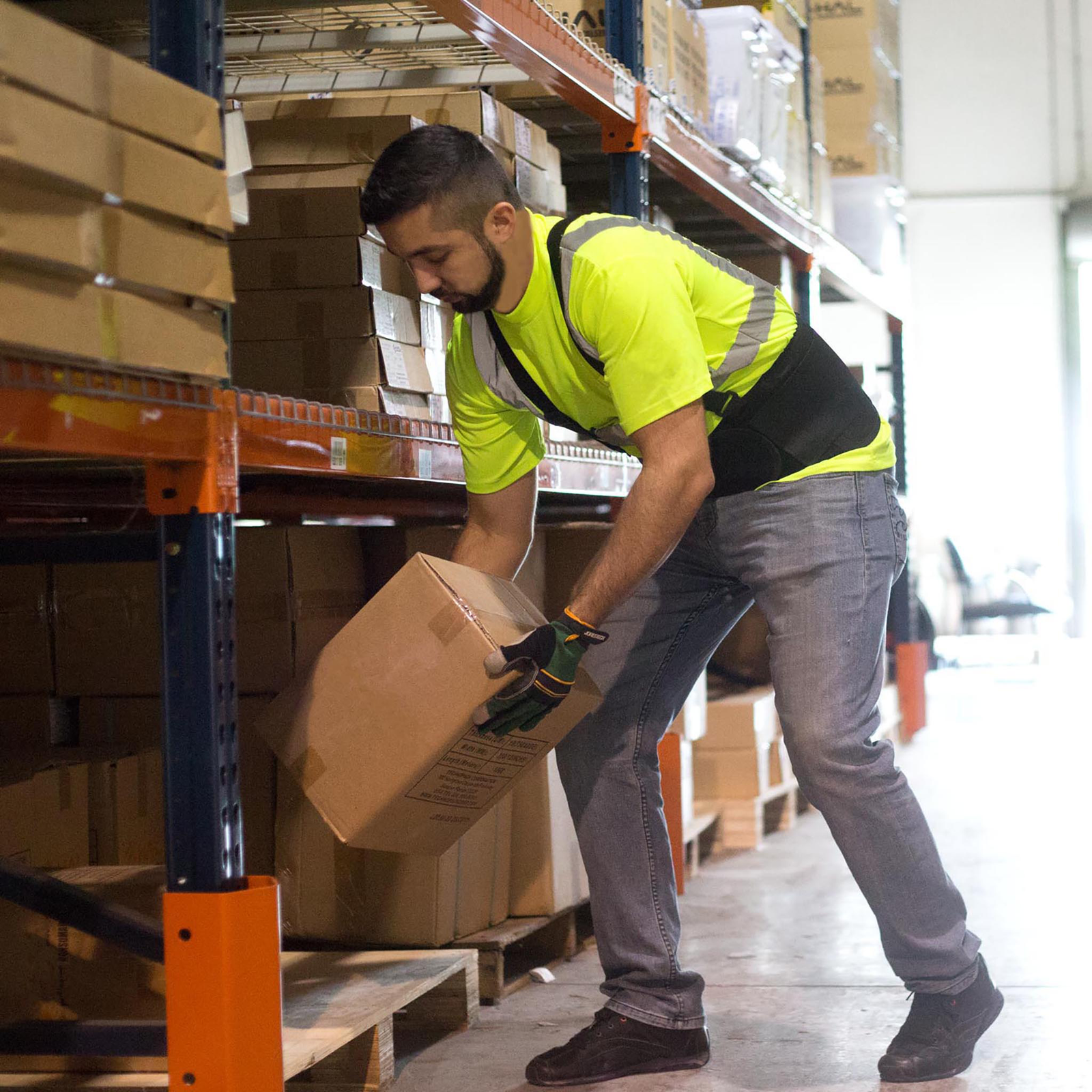 A man wearing a black adjustable back support belt with size adjusting waits straps while organizing a rack full of heavy boxes in a warehouse.
