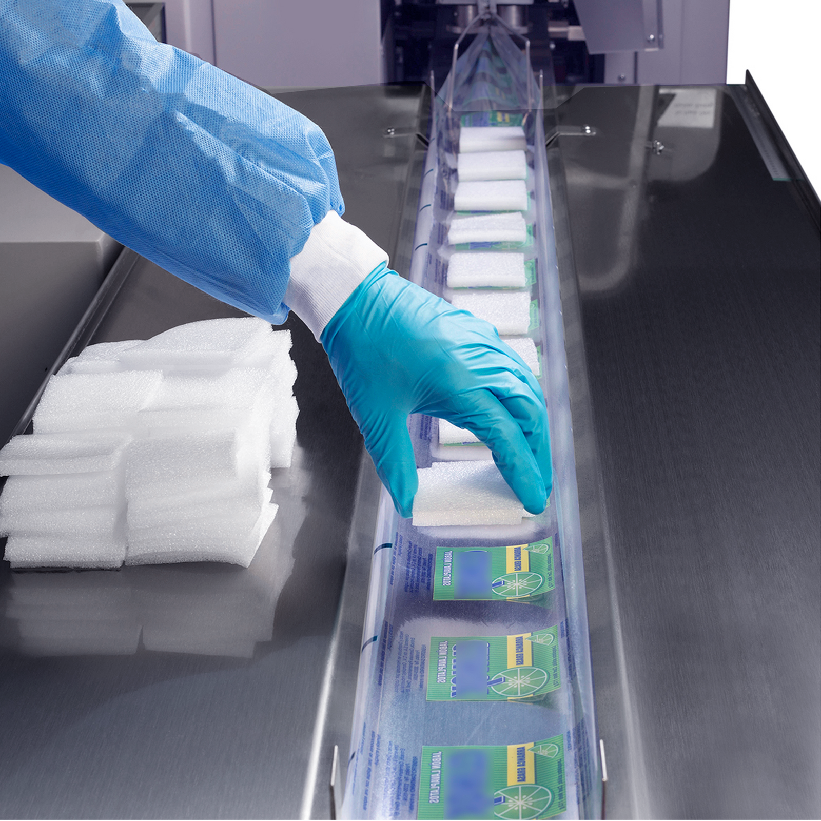 Worker operating an inverted flow pack system wearing a blue disposable glove and a blue gown. He is seen placing a white sponge-like product on top of the package film track for packaging and sealing