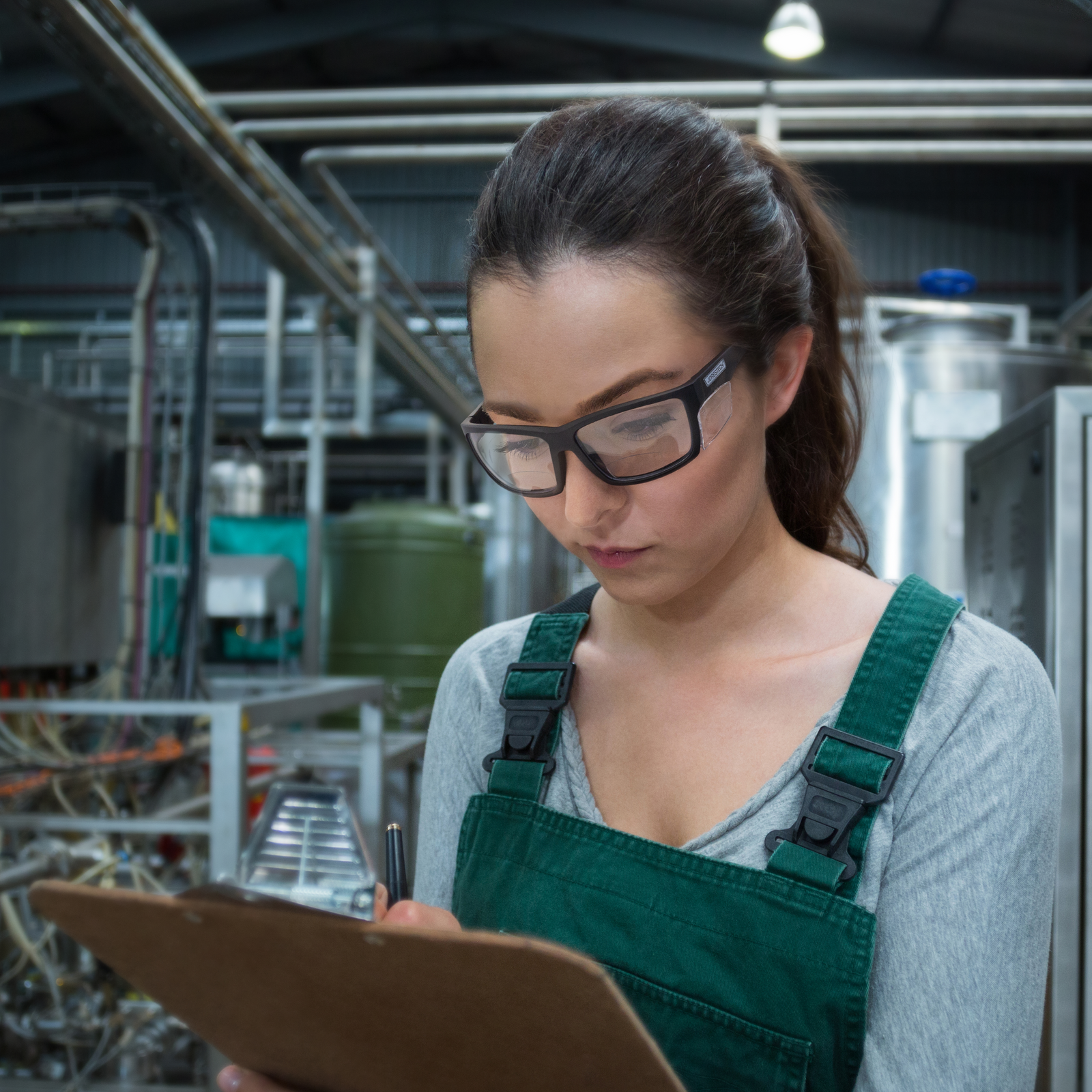 A woman working in a factory. She is wearing the bifocal JORESTECH safety reader glasses with side shields for high impact protection while taking notes on a clip board. 