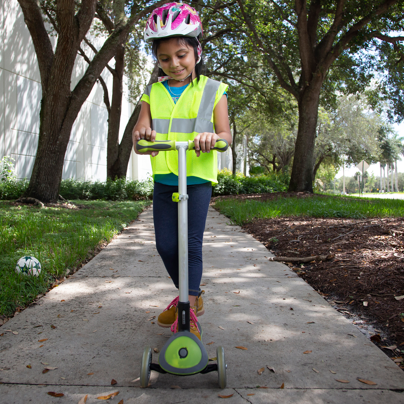 A girl wearing a safety vest for play while riding a scooter next to the road