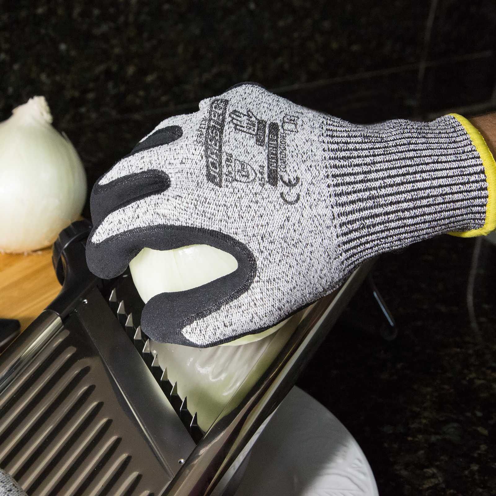 Hands of a person wearing the cut resistant JORESTECH black safety work gloves with nitrile dipped palm while cutting an onion with a silver metal blades slicer