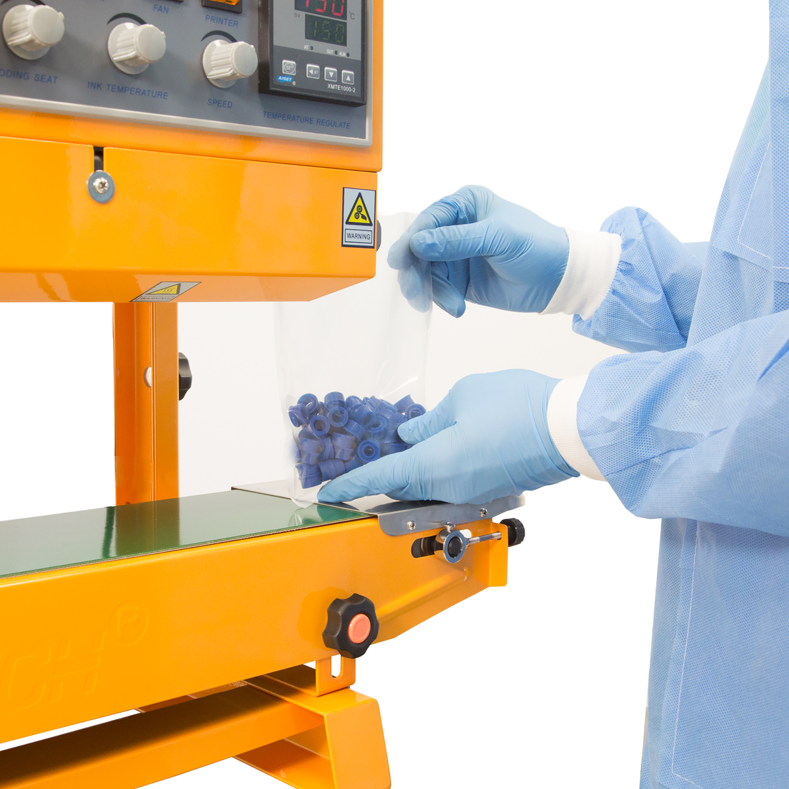 An operator wearing blue gloves and disposable clothing inserting a plastic bag filled with blue plastic parts into the yellow JORES TECHNOLOGIES® continuous band sealer. The bag sealer positioned for vertical applications