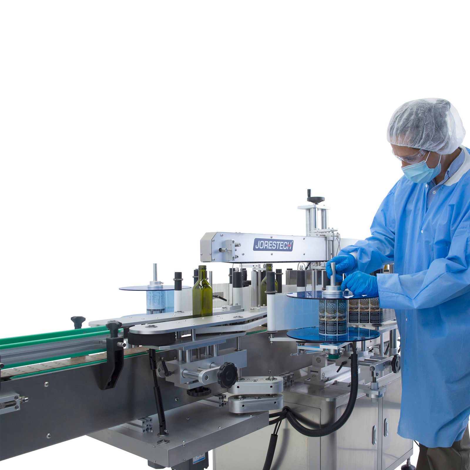 man wearing blue protective covering and hairnet adjusting knob holding the roll of labels on the JORES TECHNOLOGIES® stainless steel dual automatic label applicator for round and flat containers. Green flat glass bottles are placed on the integrated conveyor of the labeler