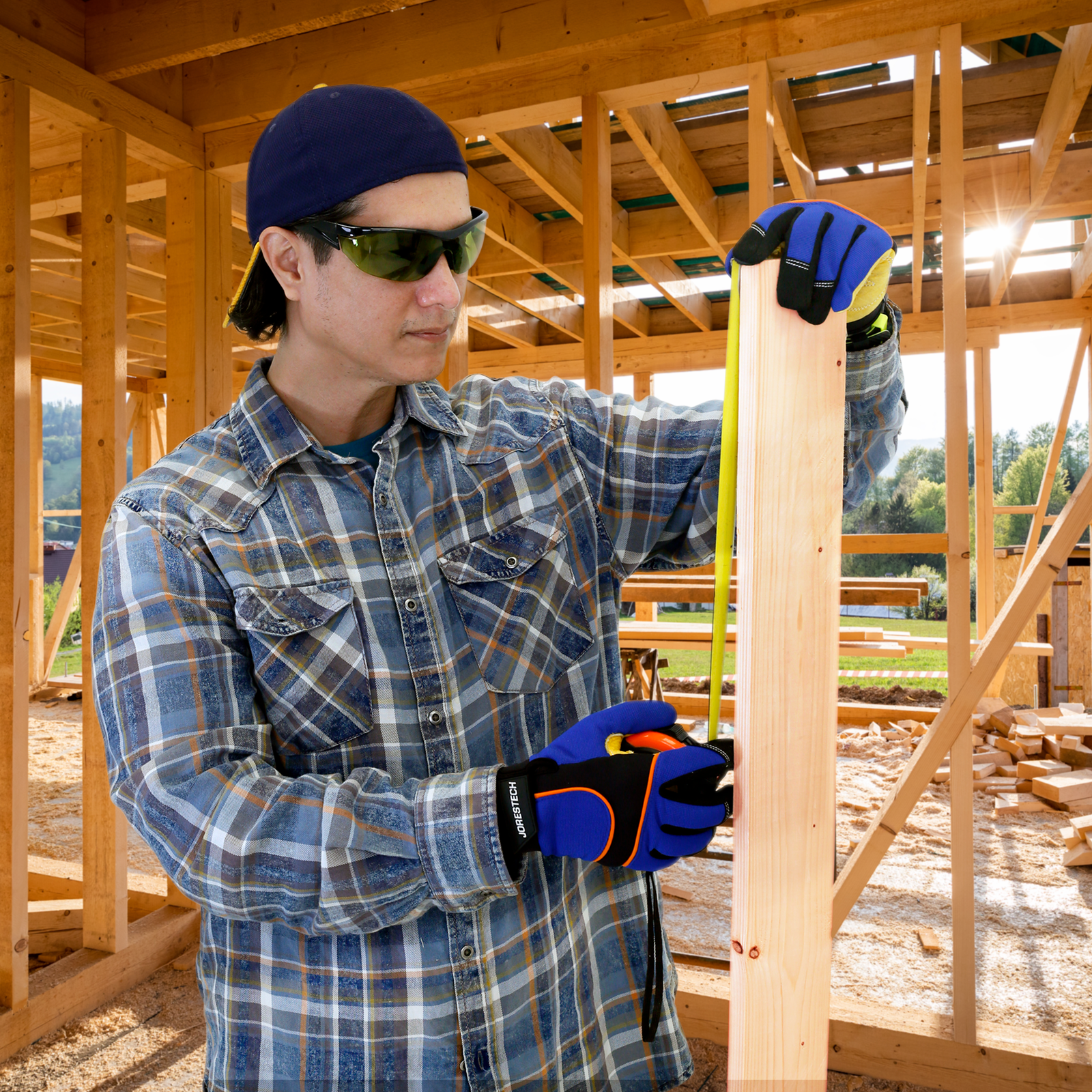 Man wearing the green framed wraparound safety glasses while measuring a 2 x 4 board to continue with the construction of a house.  The house has no walls yet so the worker uses this tinted glasses for sun protection. 