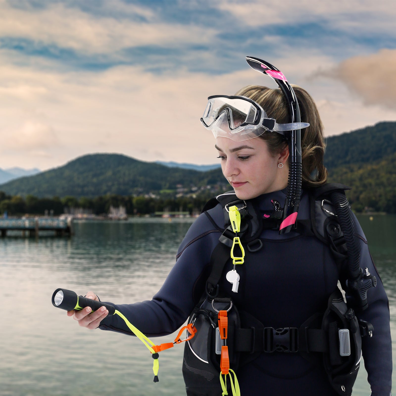 A woman wearing a black scuba gear with several glove clip safety holders of different colors attached to the buollancy control device.  Several accessories are attached to the clips like: a waterproof flash light, a safety whistle, and a mesh bag. 